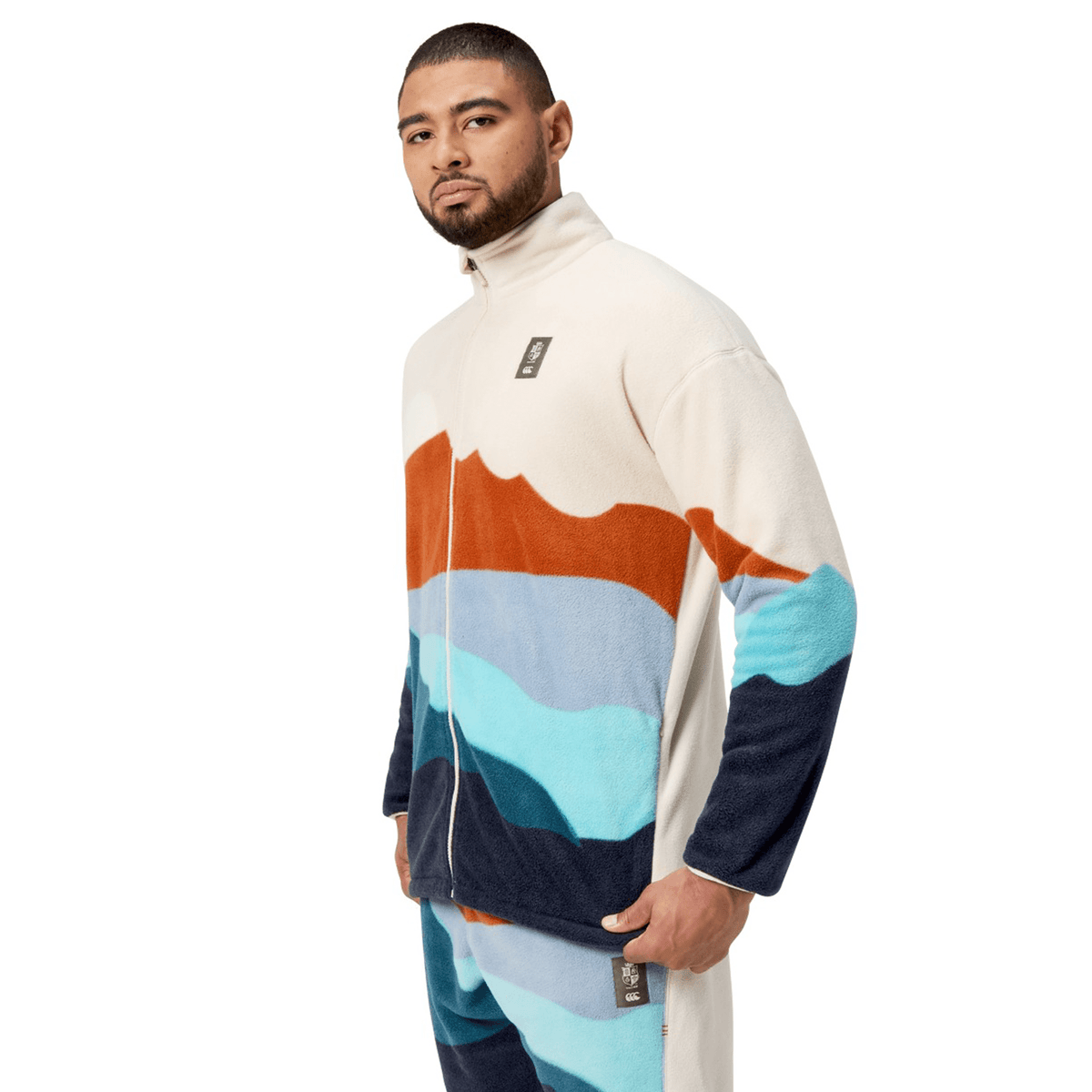 A person standing and looking forward, wearing the British &amp; Irish Lions 2025 Outback Full-Zip Fleece Jacket by Canterbury, which features a landscape design with orange, light blue, and navy hues.