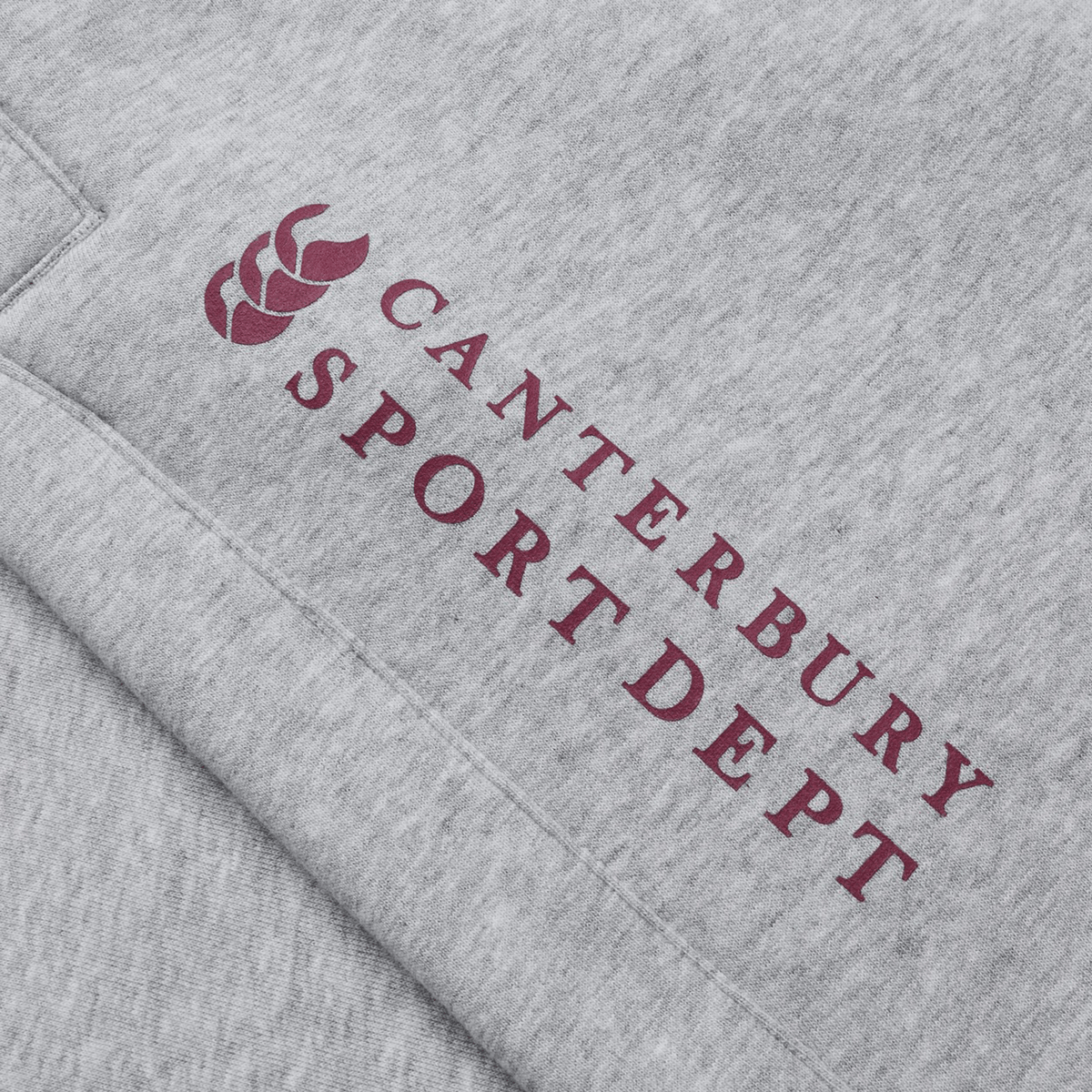 A close-up of the gray Canterbury Sport Dept Track Pant fabric features "Canterbury Sport Dept" and a maroon logo, evoking classic track pant quality, with comfort akin to an elastic waistband or cozy fleece lining.