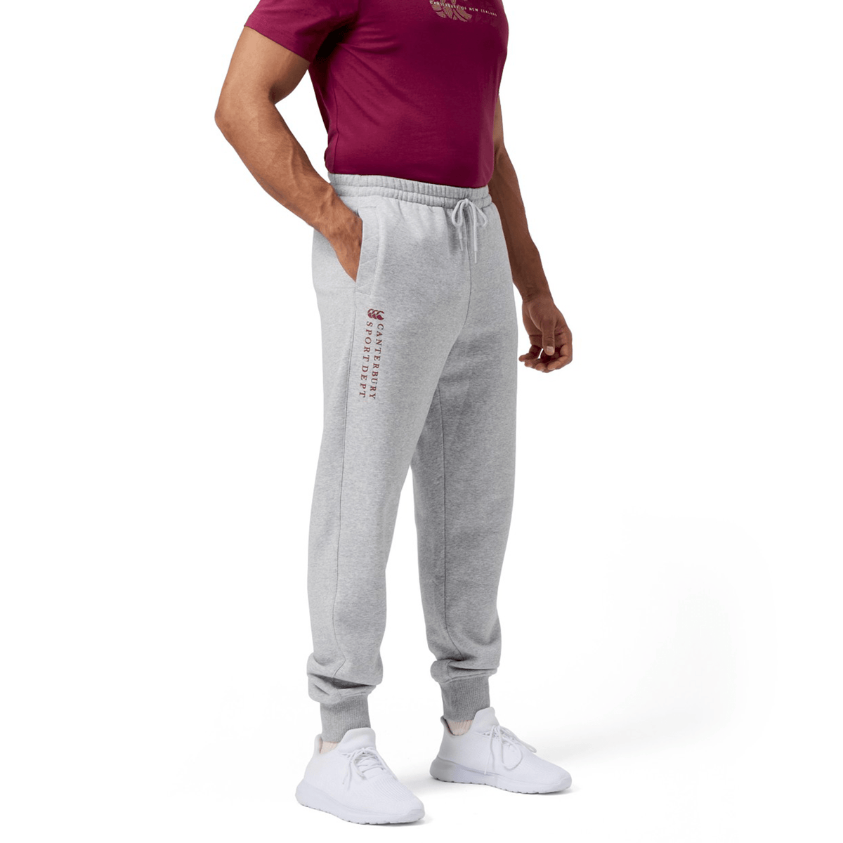 A man wearing a maroon T-shirt, Canterbury's Sport Dept Track Pant with an elastic waistband in gray, and white sneakers stands against a white background.