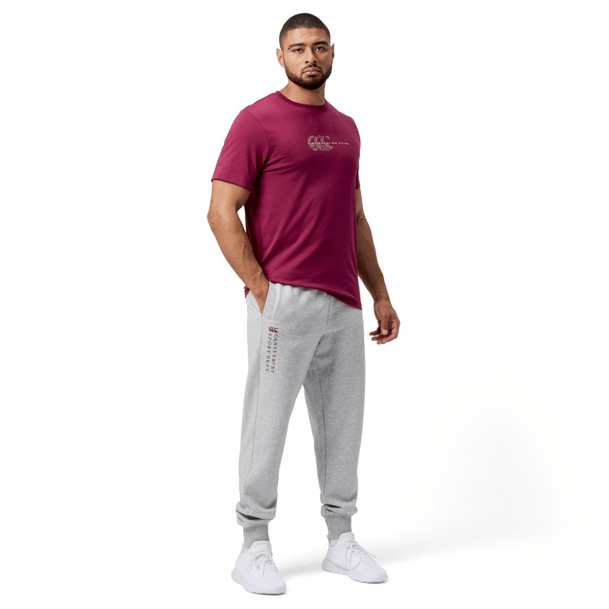 Man wearing a maroon T-shirt and Canterbury Sport Dept Track Pant by Canterbury, featuring an elastic waistband, standing with hands in pockets against a plain white background.
