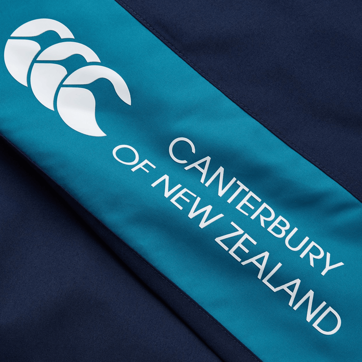 Close-up of a navy and teal fabric with Canterbury's iconic "Canterbury of New Zealand" logo featuring three stylized white "Cs," ideal for any rugby fan's collection, as seen on the Canterbury Harlequin Legends Trackpant.