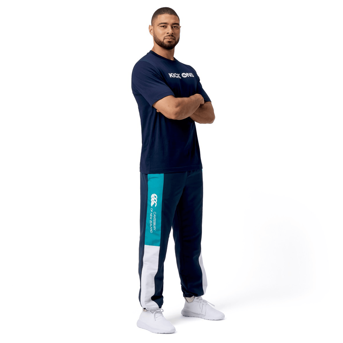 A man poses with arms crossed against a white background, wearing a navy "Kick On" T-shirt, Canterbury's Harlequin Legends Trackpant, and sneakers.