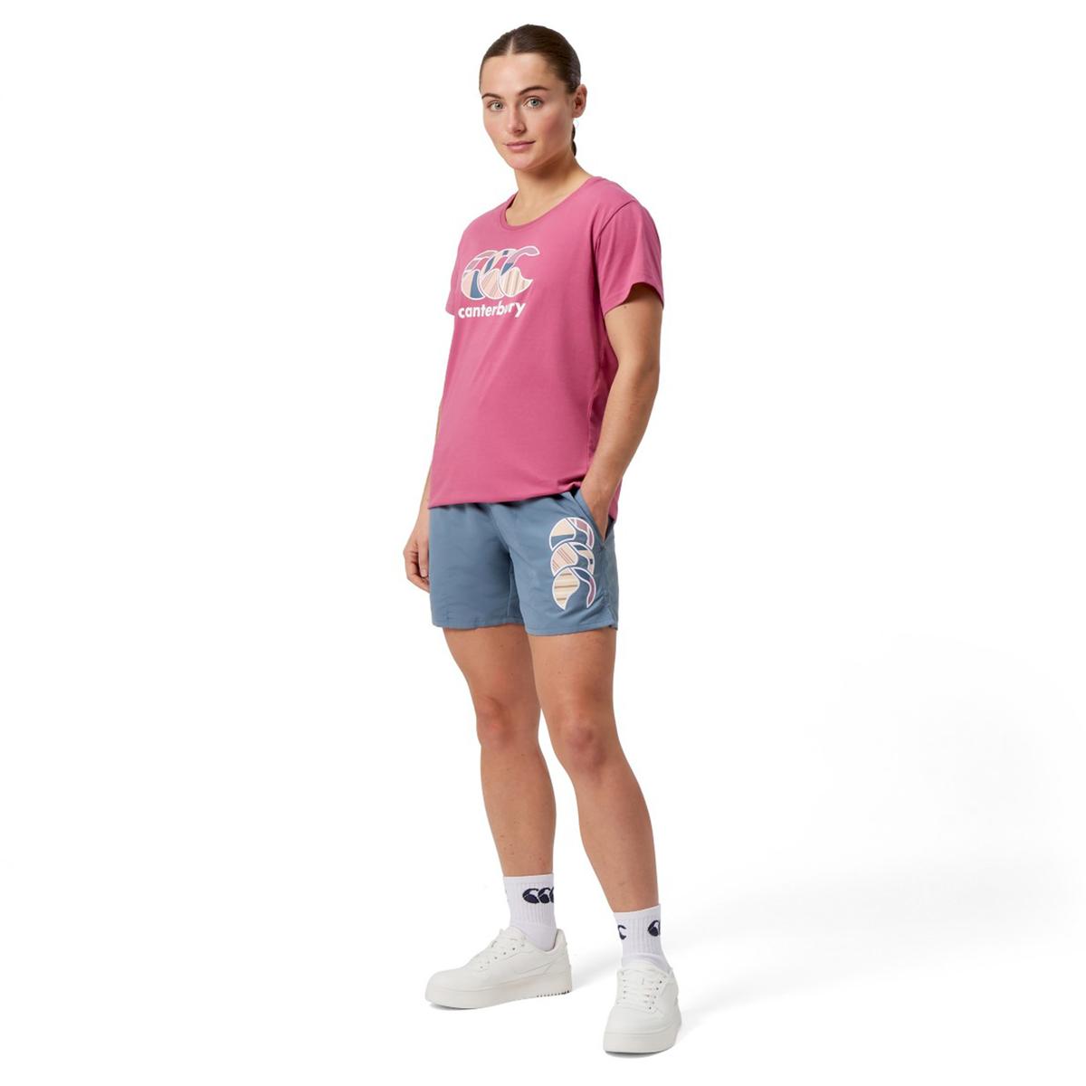 A person wearing a pink shirt and Canterbury Women&#39;s Uglies 5&quot; Tactic Short in blue, featuring a logo, stands neutrally. The look is completed with white socks and sneakers, showcasing the casual style of polyester-crafted Canterbury.