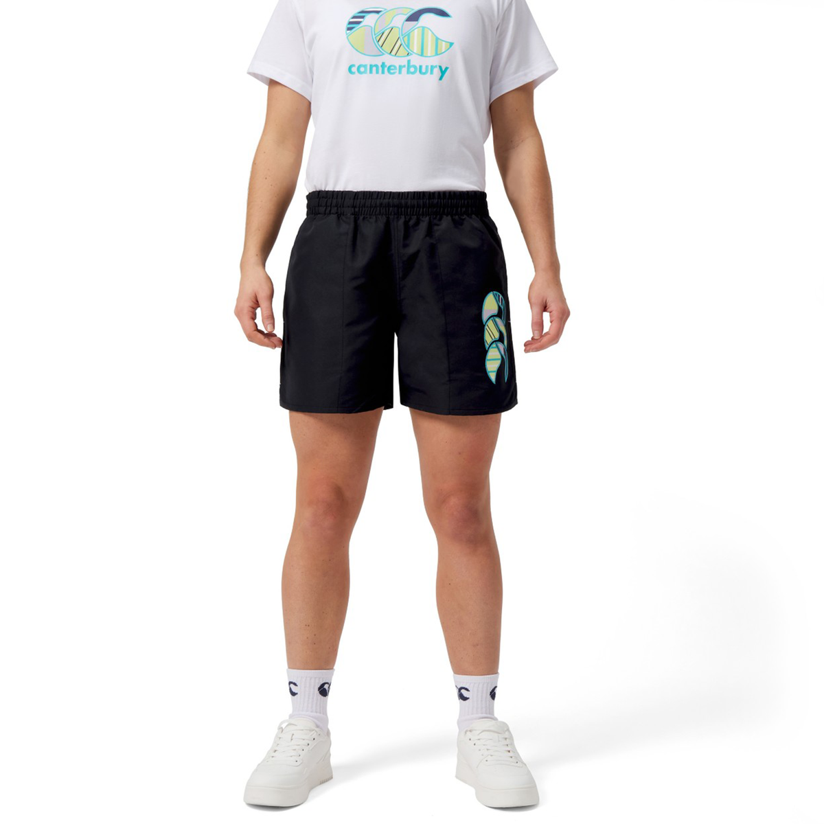 On a white background, a person wears a logo-adorned white T-shirt, black Women&#39;s Canterbury Uglies 5&quot; Tactic Shorts by Canterbury featuring vibrant Uglies emblem details, paired with white socks and sneakers.