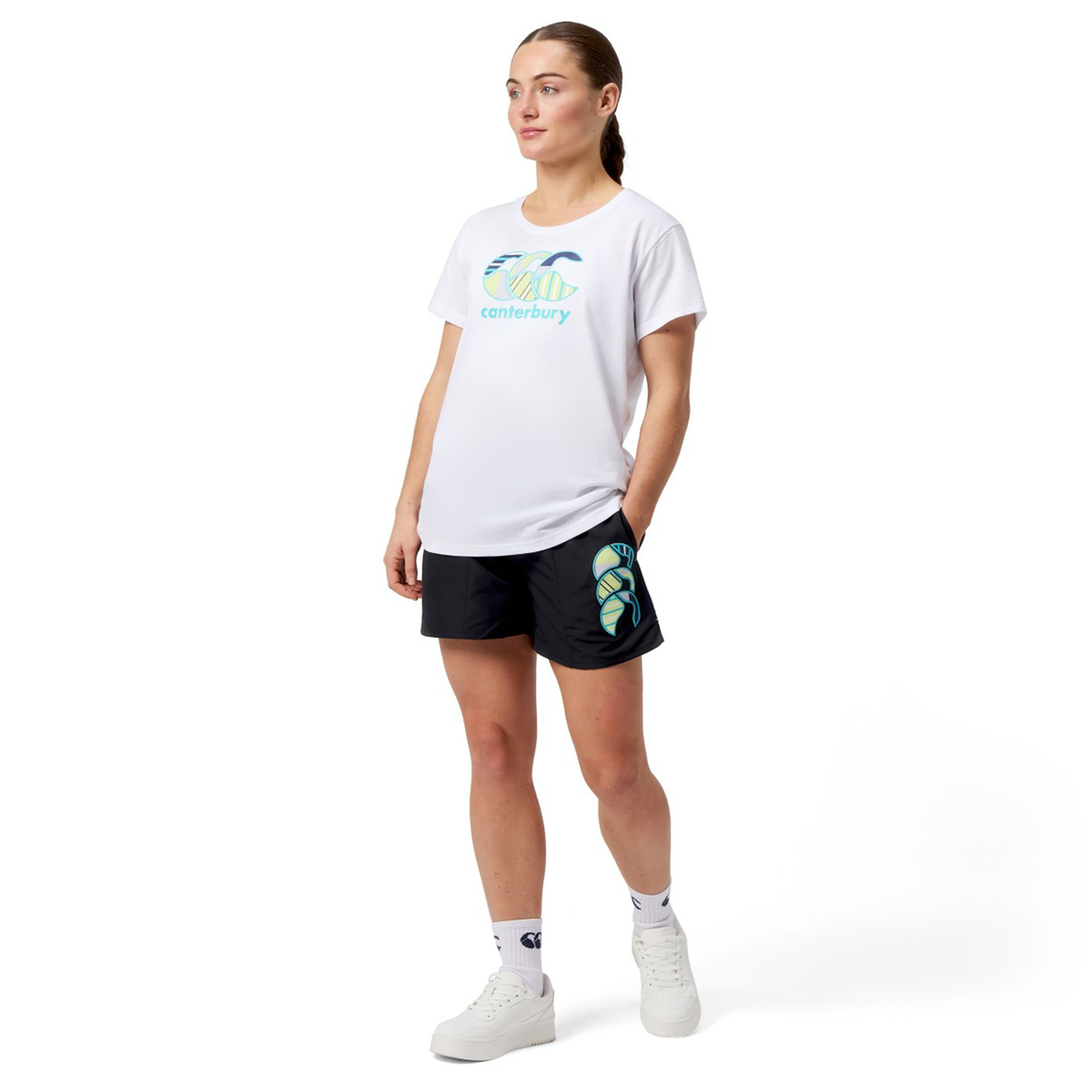 A person is standing with hands on hips, wearing a white logo T-shirt, Women&#39;s Canterbury Uglies 5&quot; Tactic Short by Canterbury in black, complemented by white sneakers and socks.