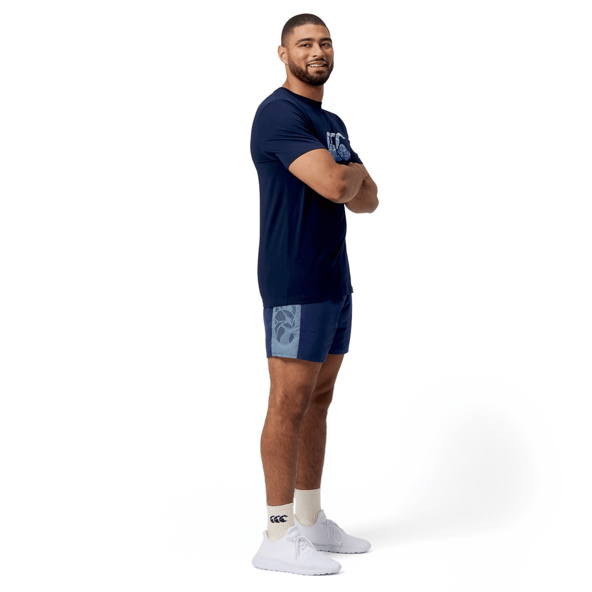 A man stands with arms crossed, wearing a navy t-shirt, Canterbury Fundamental 5" Panel Tactic Short in blue that are quick-drying, and white sneakers on a white background.