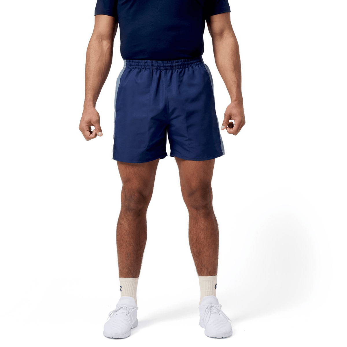 A person in a navy T-shirt, Canterbury Fundamental 5" Panel Tactic Short, and white sneakers stands against a plain white background.