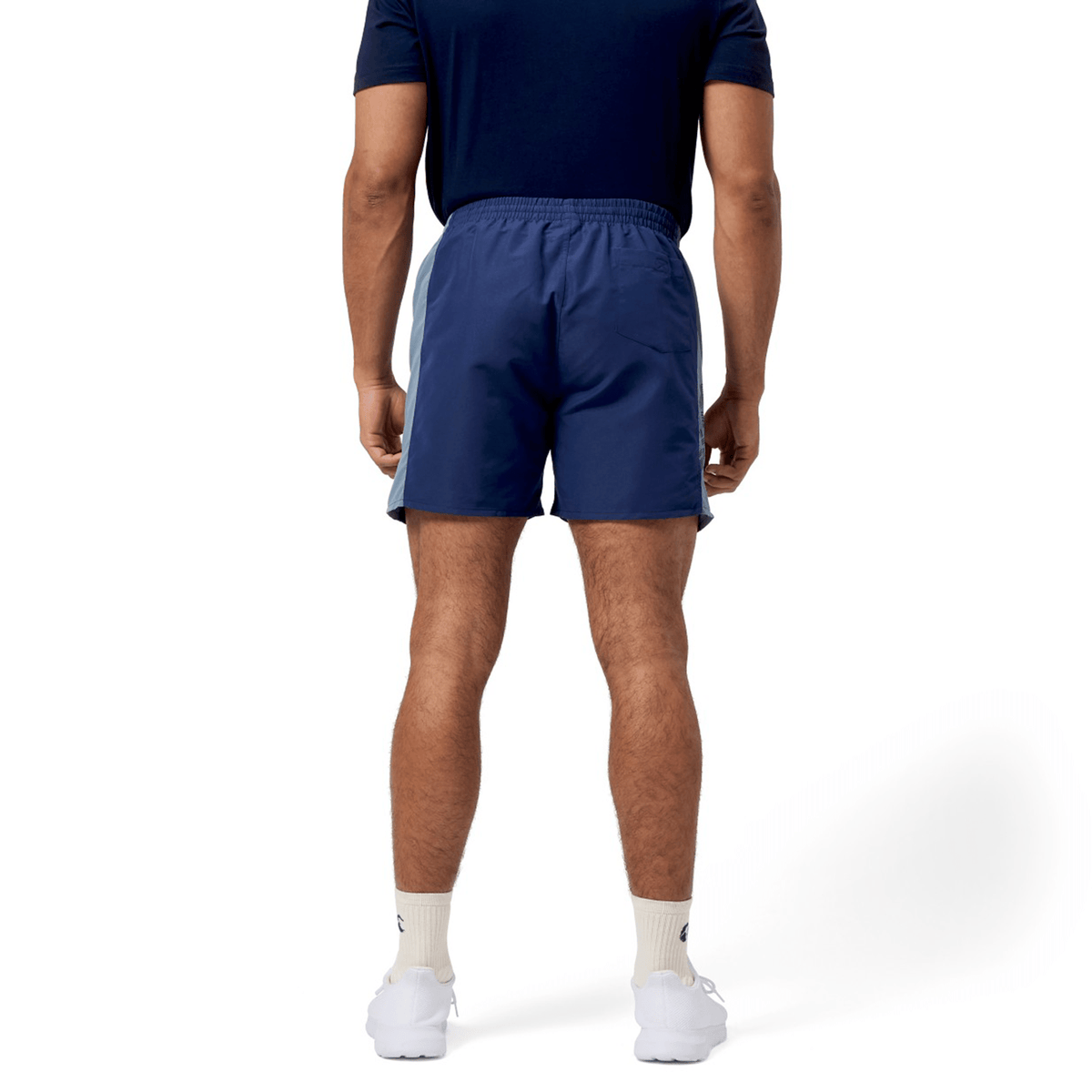A person, turning away, sports a navy shirt and Canterbury Fundamental 5" Panel Tactic Short. Against a white backdrop, their white socks and sneakers pop. The moisture-wicking shorts by Canterbury promise quick drying for any activity.