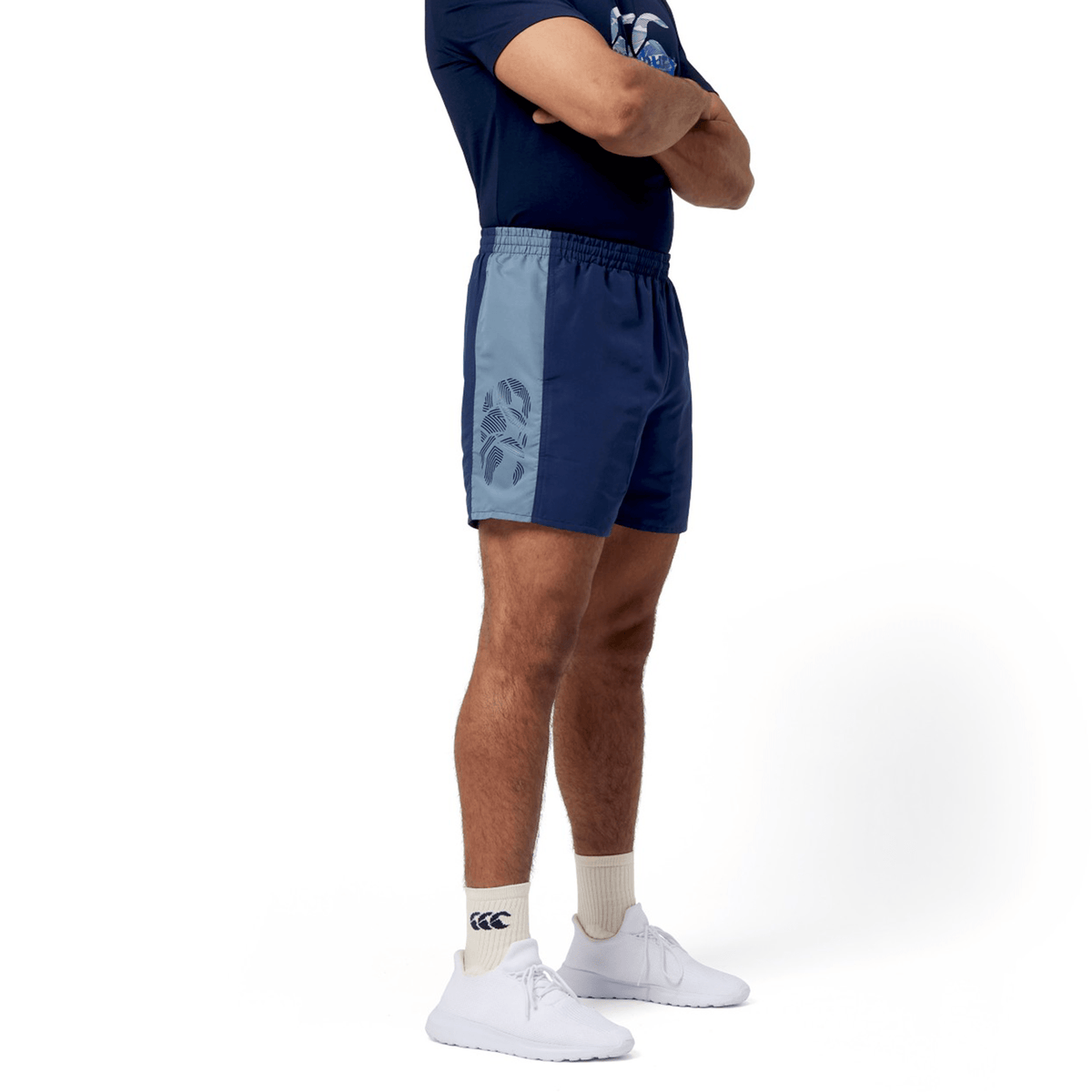 A man in athletic attire confidently stands with arms crossed, wearing a navy and light blue outfit featuring the Canterbury Fundamental 5" Panel Tactic Short. He completes his look with white sneakers and logo-adorned socks, ideal for those seeking style and performance in quick-drying shorts.