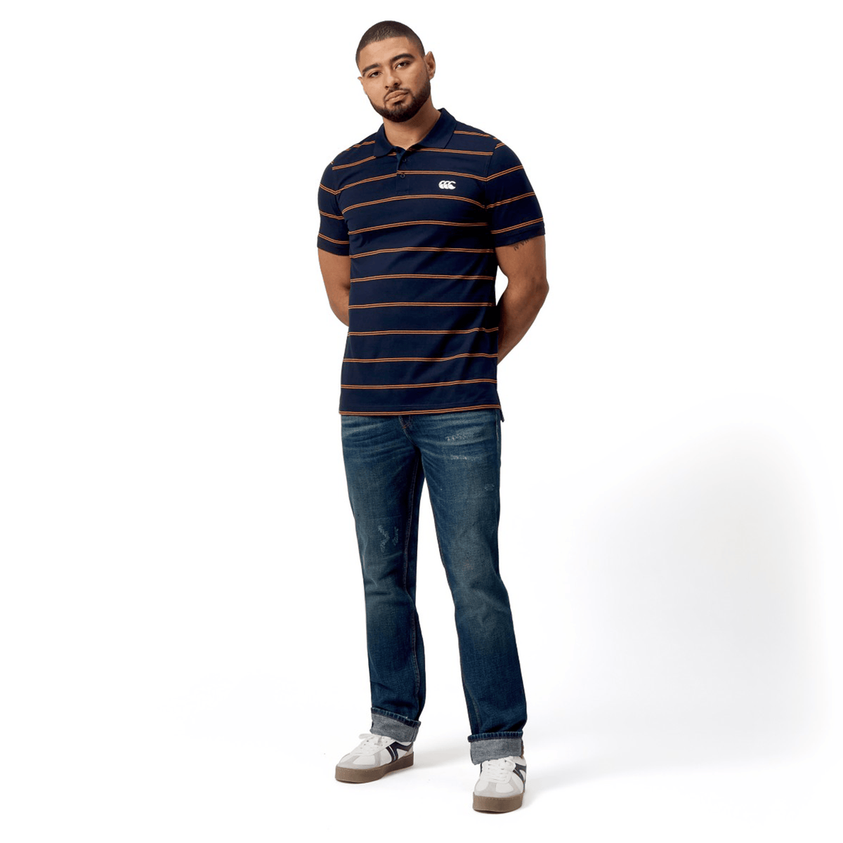 A person in a Canterbury Fundamental Yarn-Dye Stripe Polo, blue jeans, and white sneakers stands against a plain Canterbury white backdrop.