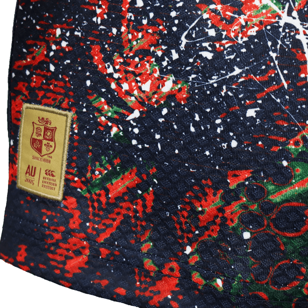 Close-up of the Youth British &amp; Irish Lions 2025 Warm Up Jersey by Canterbury, showcasing moisture-wicking fabric featuring a vibrant abstract design in red, green, and white on a dark background, with a beige patch resembling the UK Lions crest and &quot;AU&quot; and &quot;2003&quot; text.