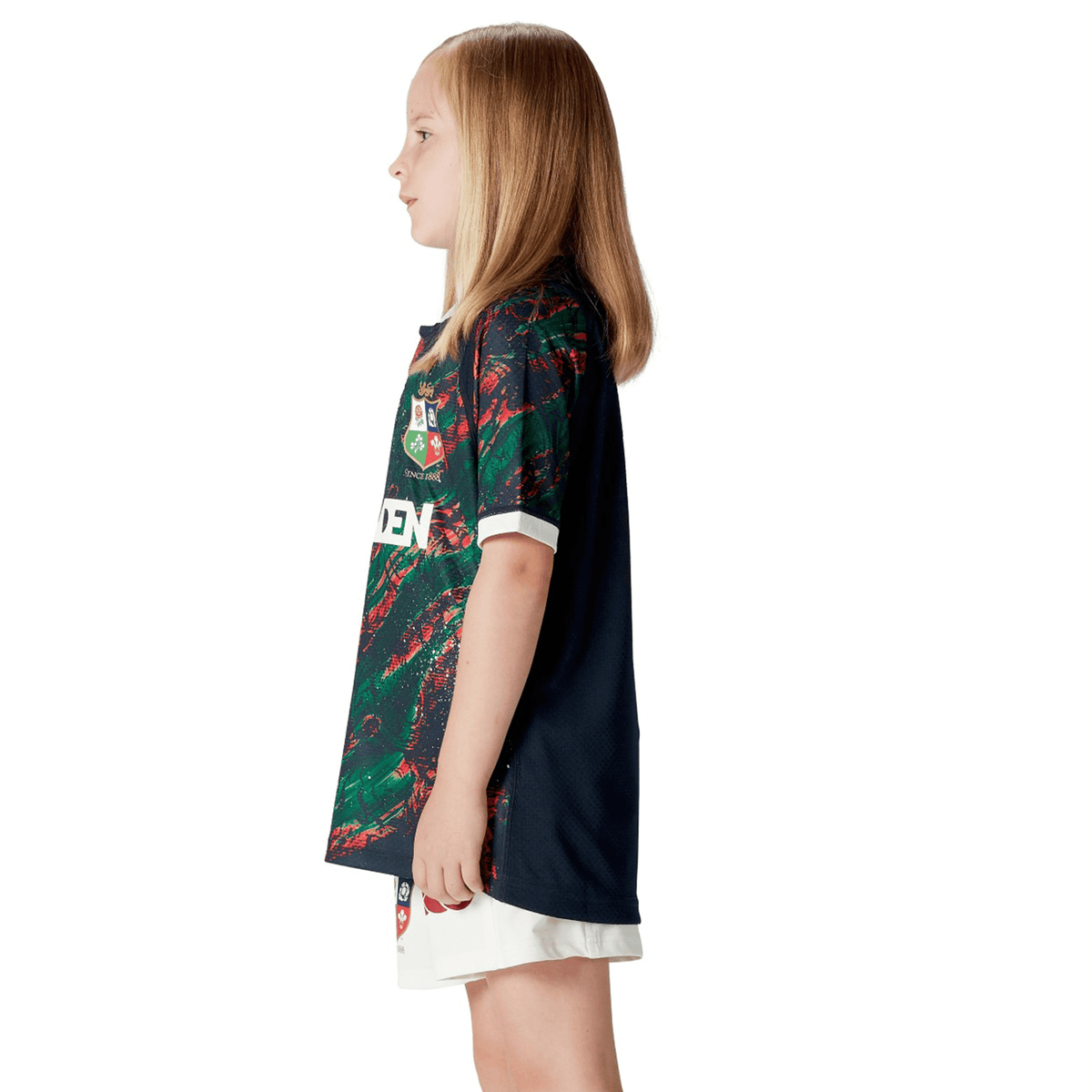 A young girl with long hair stands in profile, proudly wearing the Youth British &amp; Irish Lions 2025 Warm Up Jersey by Canterbury, crafted from moisture-wicking fabric, paired with crisp white shorts.