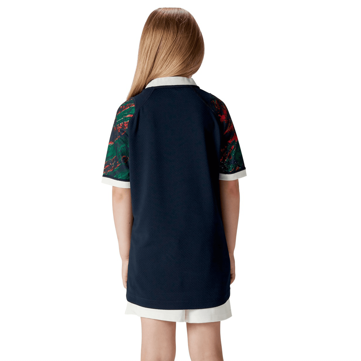 From behind, a child with flowing hair is seen wearing the Youth British &amp; Irish Lions 2025 Warm Up Jersey by Canterbury, featuring a navy-blue color, short sleeves, crisp white collar, and patterned details.