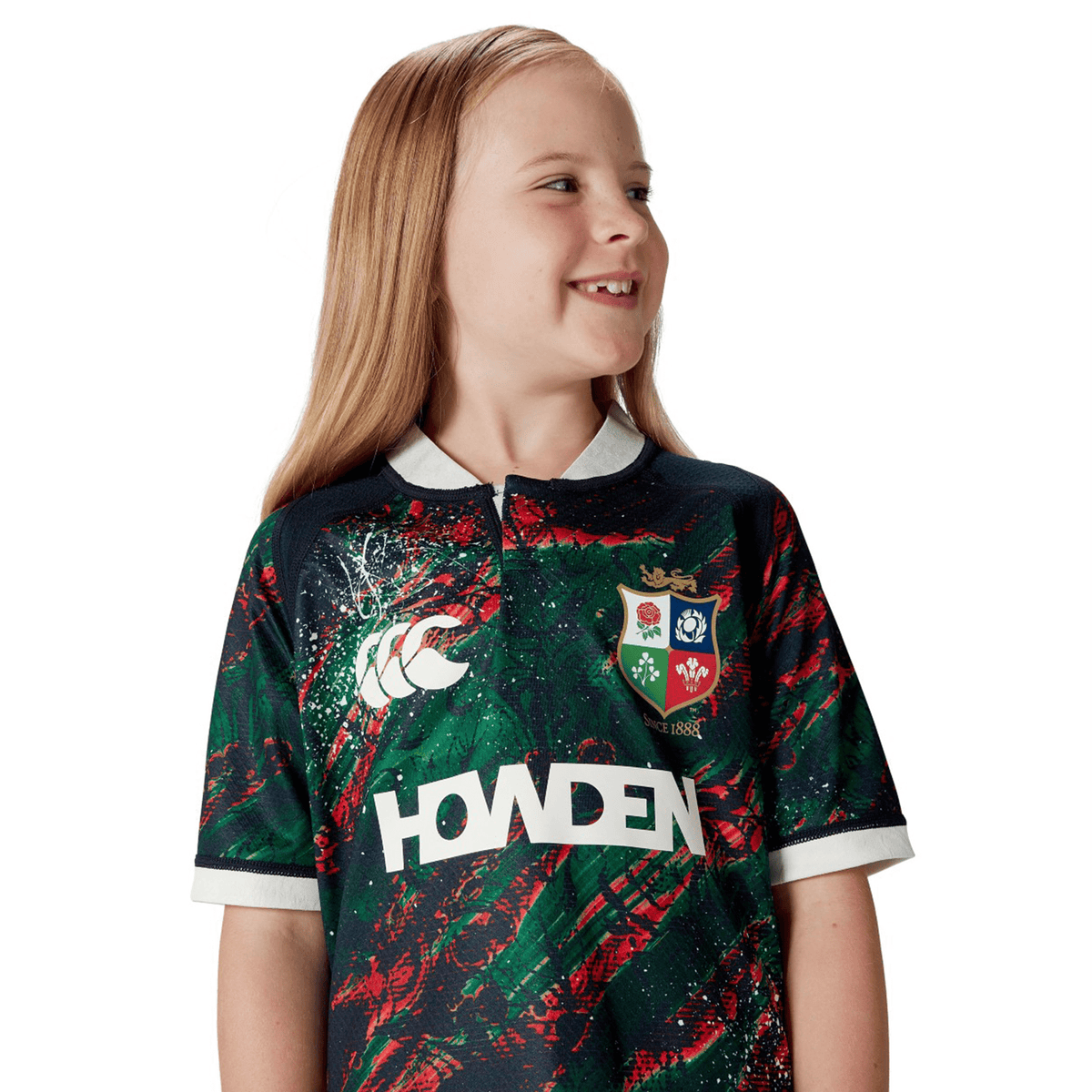 A child smiles while wearing the Youth British &amp; Irish Lions 2025 Warm Up Jersey by Canterbury, glancing to the side against a plain white backdrop. The jersey&#39;s moisture-wicking fabric ensures comfort, making it ideal for young Lions fans.