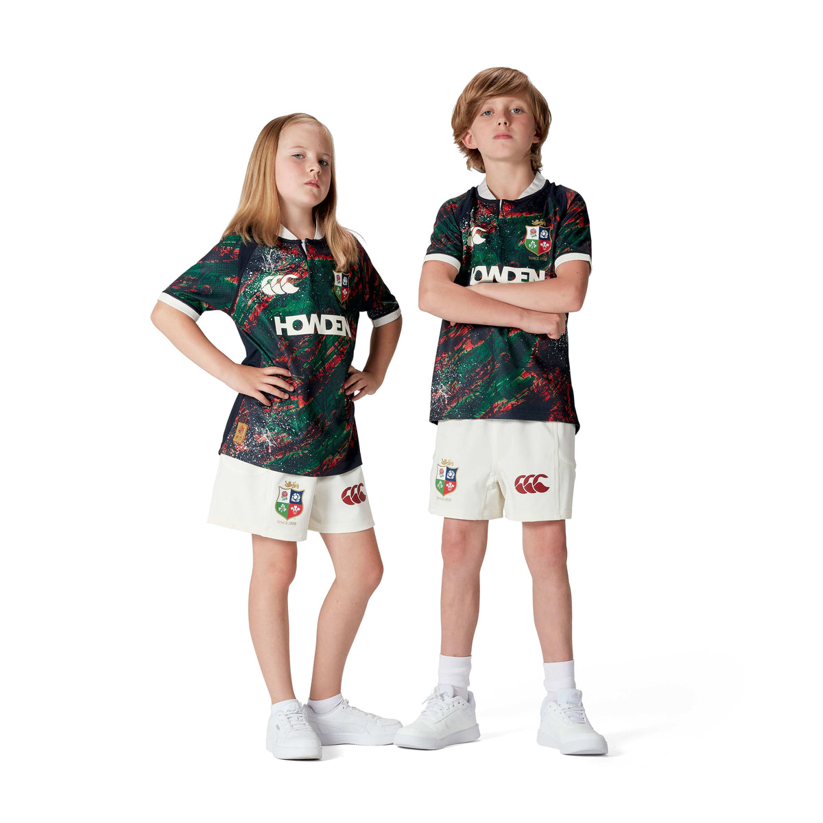 Two kids stand confidently in matching sports uniforms with a green and red abstract pattern and white shorts. Made from moisture-wicking fabric, these outfits echo the style of the Youth British &amp; Irish Lions 2025 Warm Up Jersey by Canterbury against a clean, white backdrop.