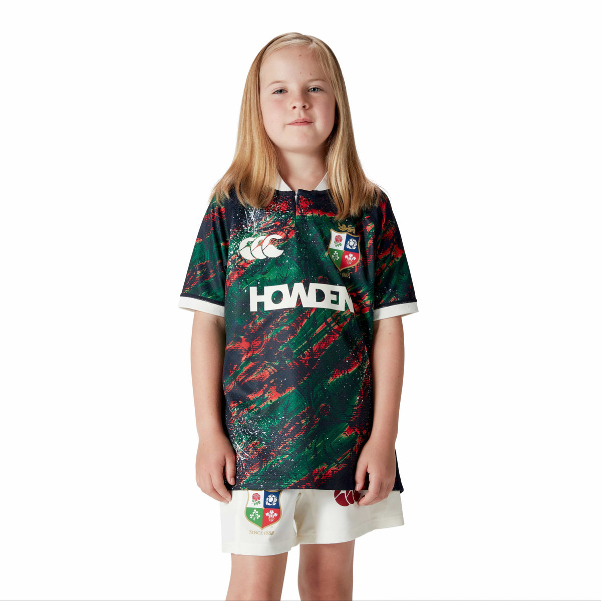 A child wearing the Canterbury Youth British &amp; Irish Lions 2025 Warm Up Jersey and white shorts poses against a plain white background, highlighting its vibrant design.
