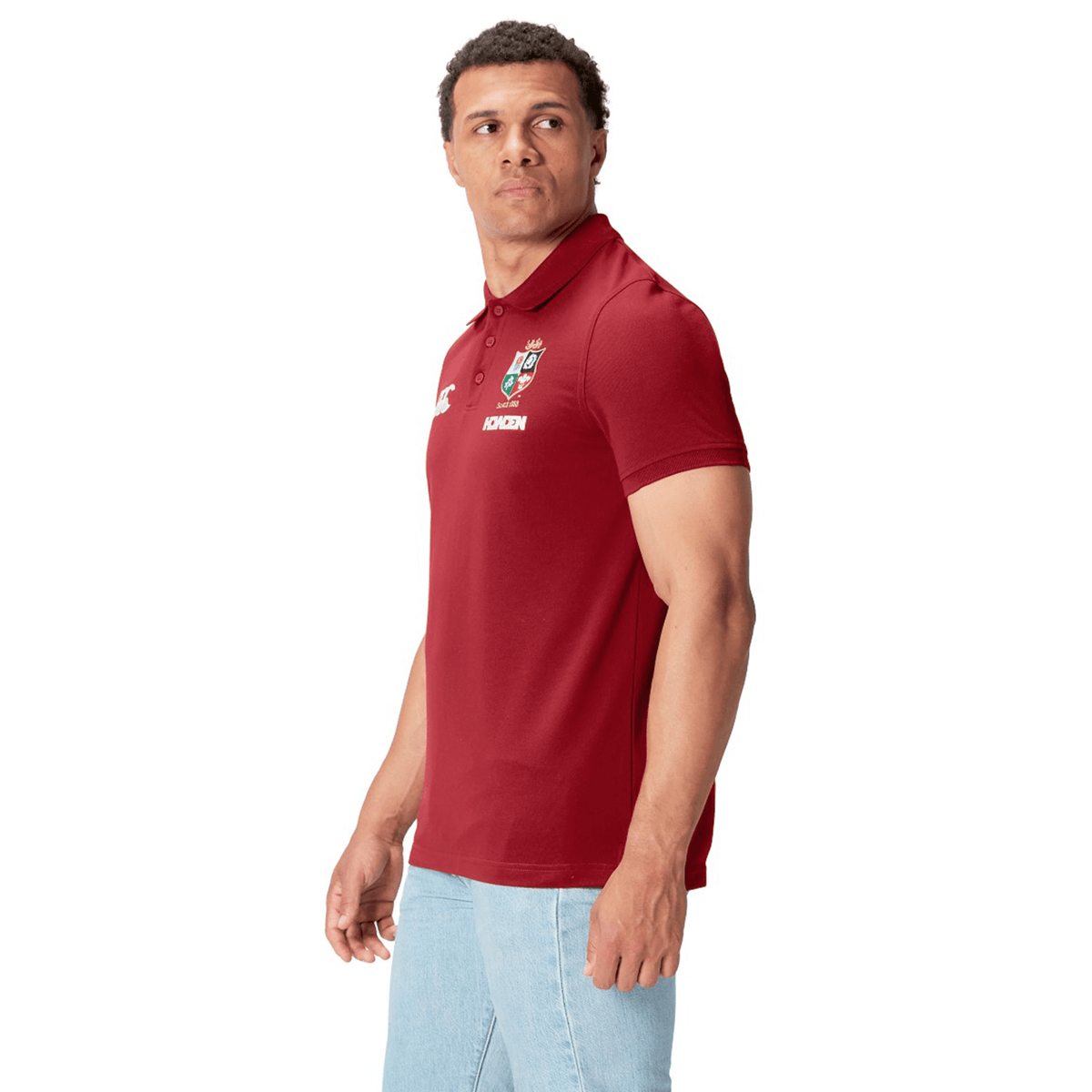 A person wears a British &amp; Irish Lions 25 Pique Polo by Canterbury in red with a white logo, echoing the classic Canterbury style, paired with light blue jeans. They gaze to the side against a plain white background.