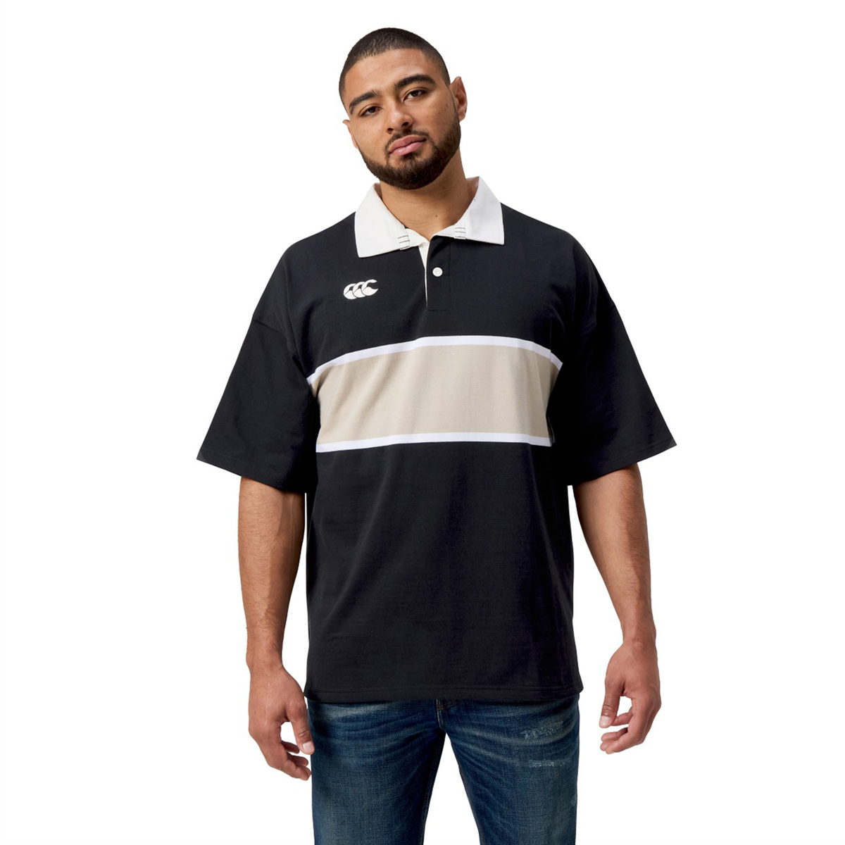 A person stands against a white background, wearing the Canterbury Yarn Dye Short Sleeve Rugby Jersey, featuring a black base with a distinctive beige and white stripe across the chest.