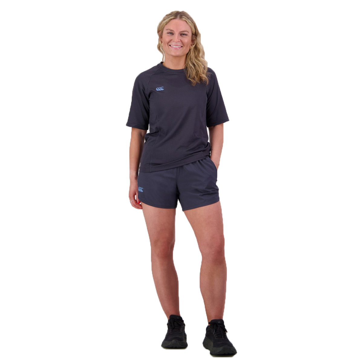 A person in a dark athletic outfit stands smiling against a white background, showcasing the Women&#39;s Canterbury Tempo+ 5&quot; Training Short, made from a comfortable polyester-elastane blend.