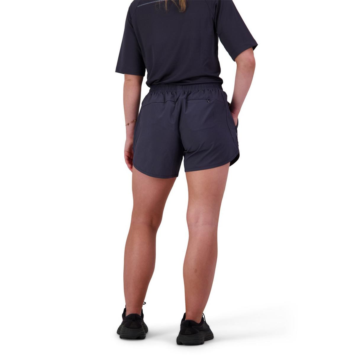 Facing away, a person wears a dark athletic shirt and Women&#39;s Canterbury Tempo+ 5&quot; Training Short made from a polyester-elastane blend. The look is complemented by black sneakers, with reflective piping subtly visible against the white background and hands casually in pockets.