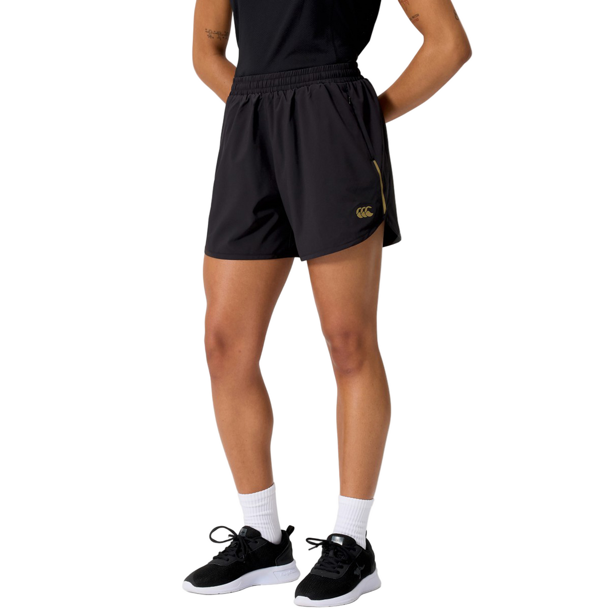 A person wearing a black t-shirt, Canterbury Women&#39;s Tempo+ 5&quot; Training Shorts, white socks, and black sneakers stands against a white background.