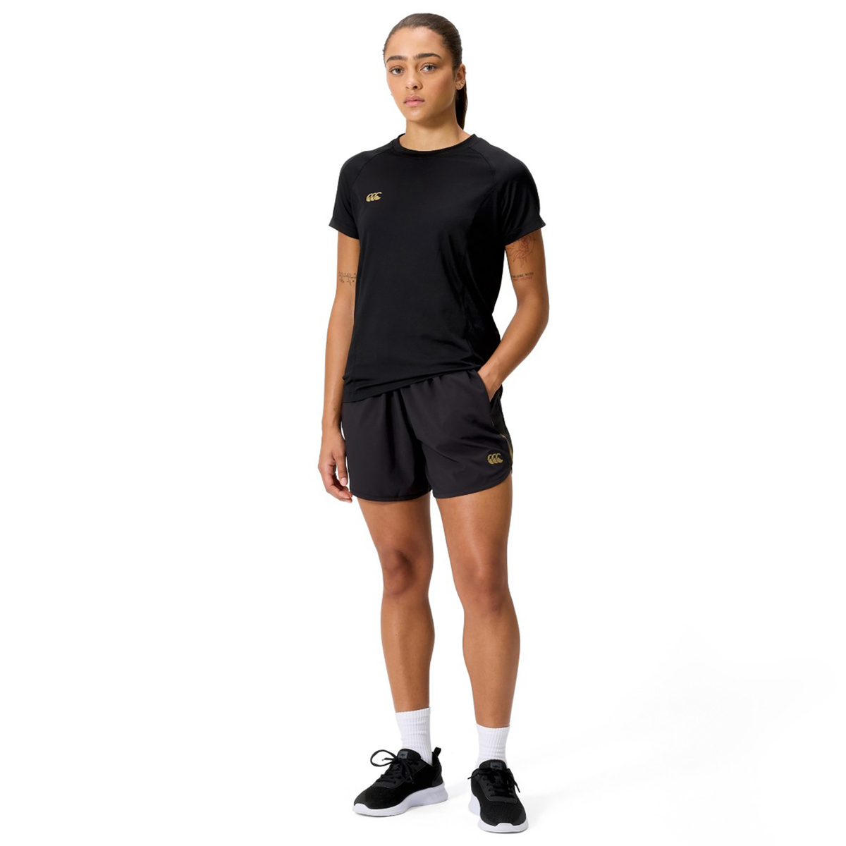 A person faces forward wearing a black t-shirt, Canterbury Women&#39;s Tempo+ 5&quot; Training Shorts in black, white socks, and black sneakers against a plain white background.