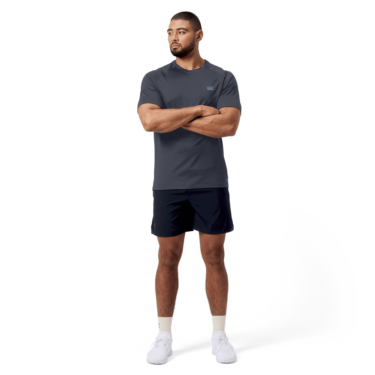 A man stands with arms crossed, exuding a dynamic look in a Canterbury Tempo+ Training Tee and black moisture-wicking shorts. He pairs these with light socks and white shoes against a plain white background.