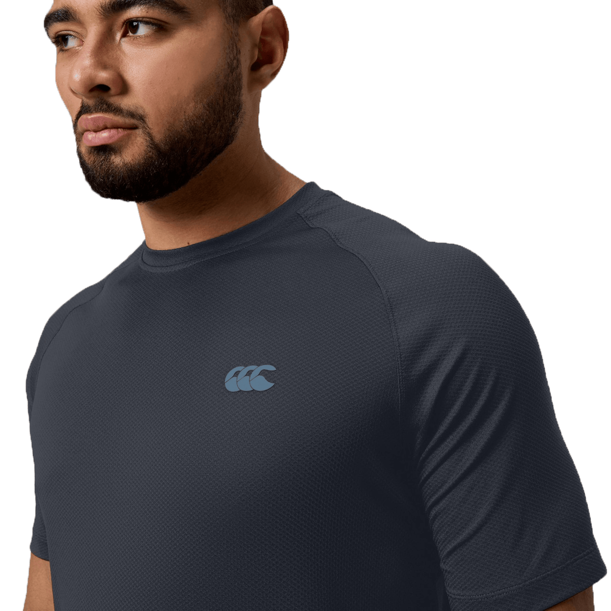 A man in a navy Canterbury Tempo+ Training Tee with a blue sports logo on the chest looks to the side. The dynamic look is enhanced by moisture-wicking fabric, ensuring comfort and style.