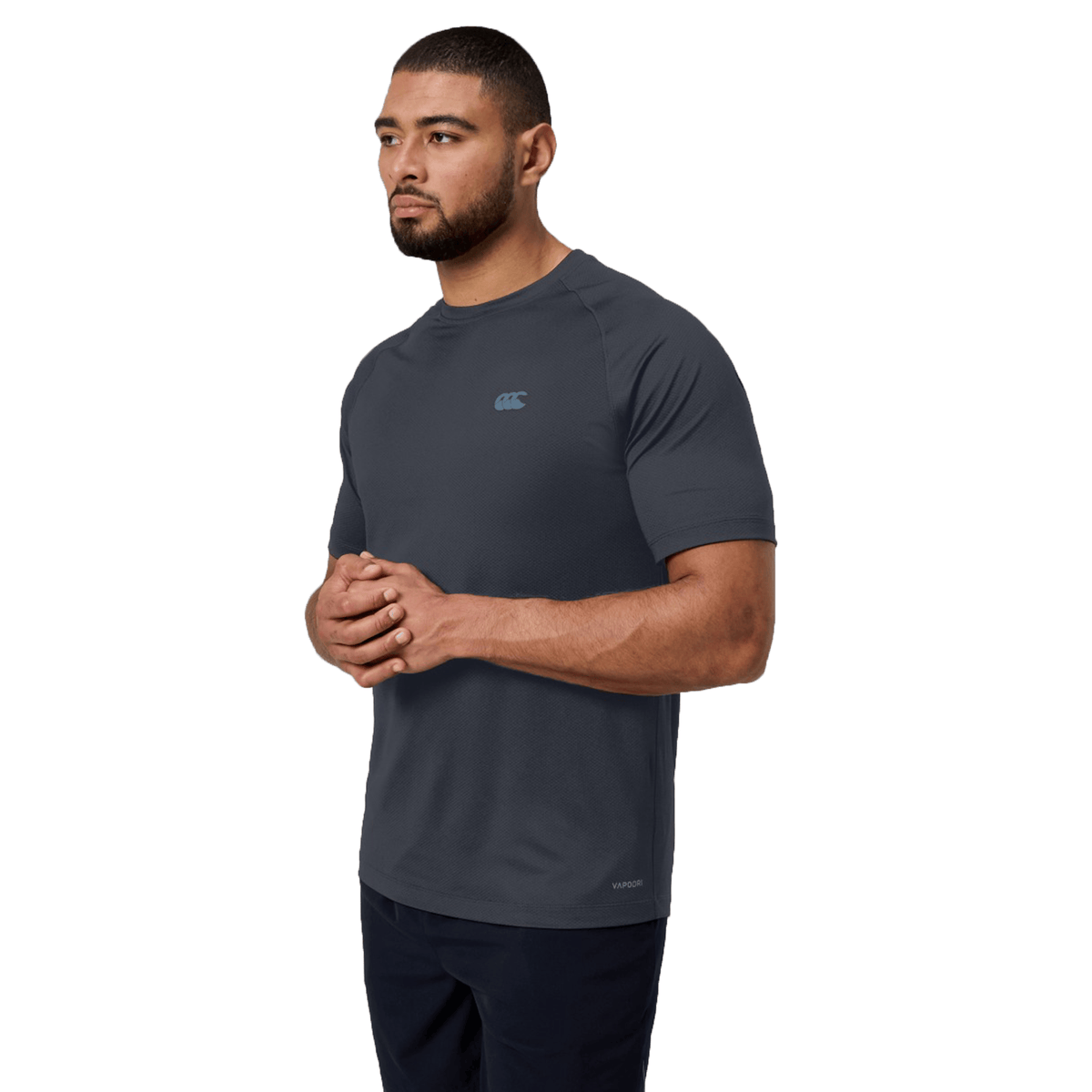 A man wears the dark gray Canterbury Tempo+ Training Tee, featuring a white logo and moisture-wicking fabric, standing with hands clasped. With a neutral expression and facing slightly to the side, the plain white background highlights the shirt's dynamic look.