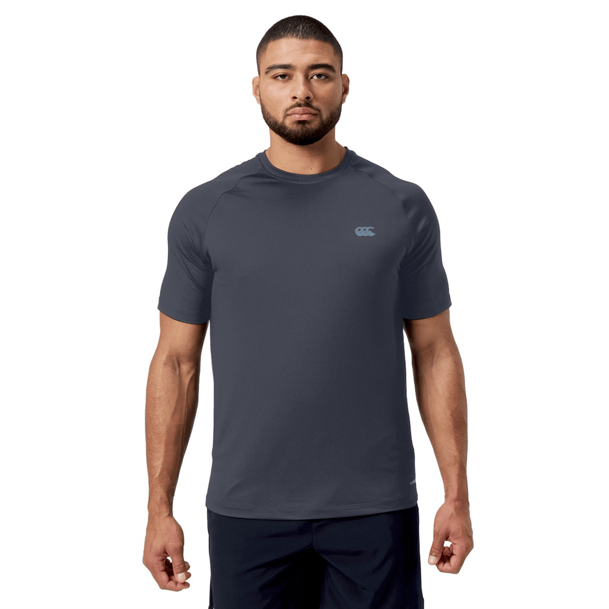 A man with short hair and a beard, showcasing a dynamic style, wears the Canterbury Tempo+ Training Tee in dark gray alongside black shorts, all set against a white background. The T-shirt is made from moisture-wicking fabric for comfort and performance.