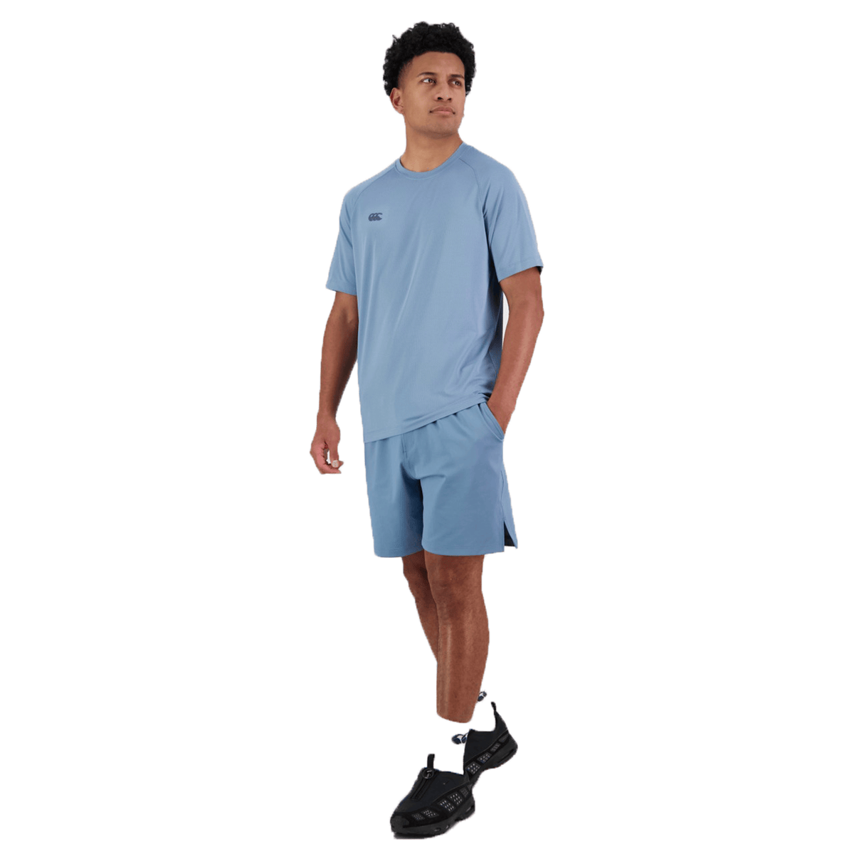 A person in a blue Canterbury Tempo+ Training Tee, featuring moisture-wicking fabric, and black sneakers stands against a white background with one hand in their pocket.