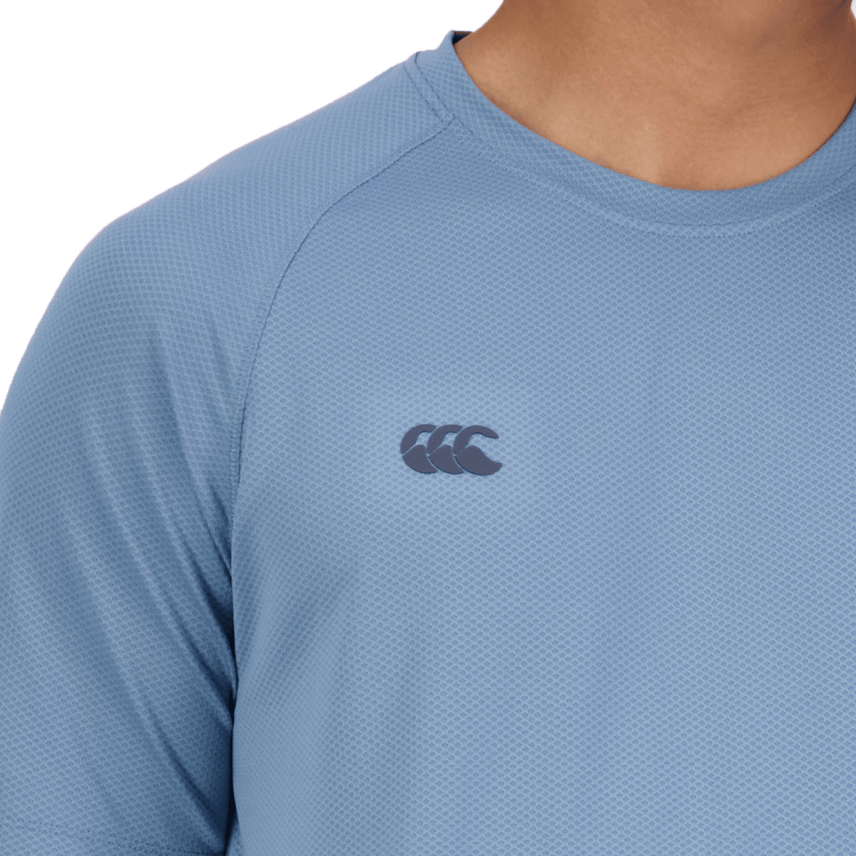 A glimpse of someone in a light blue Canterbury Tempo+ Training Tee shows a small dark logo with three overlapping curves on the chest, made from moisture-wicking fabric.