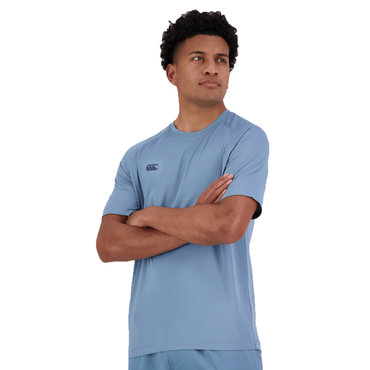 A person in a Canterbury Tempo+ Training Tee, a light blue athletic outfit made from moisture-wicking fabric, stands with arms crossed, gazing to the side against a white background.