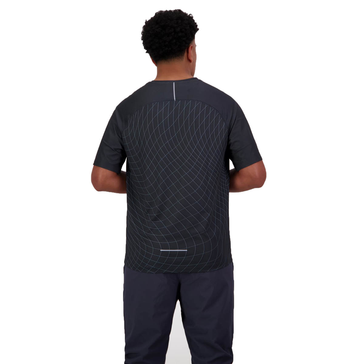 A person with short curly hair stands facing away, wearing a Canterbury Tempo+ AOP Training Tee in black, paired with dark pants. The tee features a subtle geometric pattern and moisture-wicking fabric, providing comfort and style for any activity.