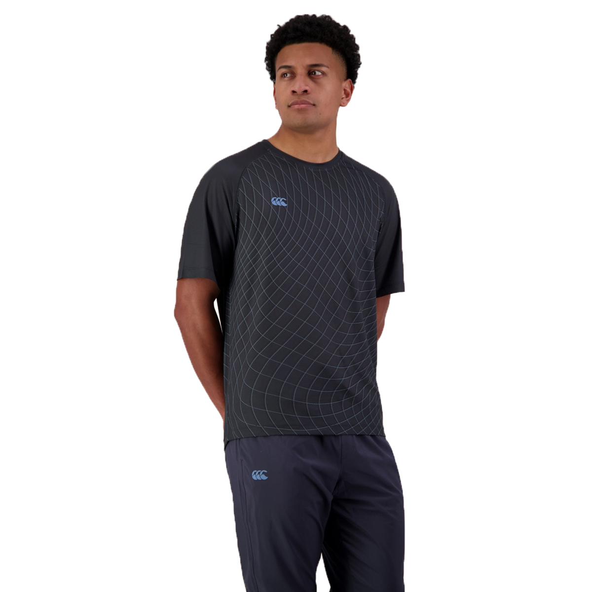 Against a white background, a person wears the Canterbury Tempo+ AOP Training Tee, featuring a dark geometric design made of moisture-wicking fabric. The outfit, paired with dark pants and their hands casually behind their back, is ideal for performance training.