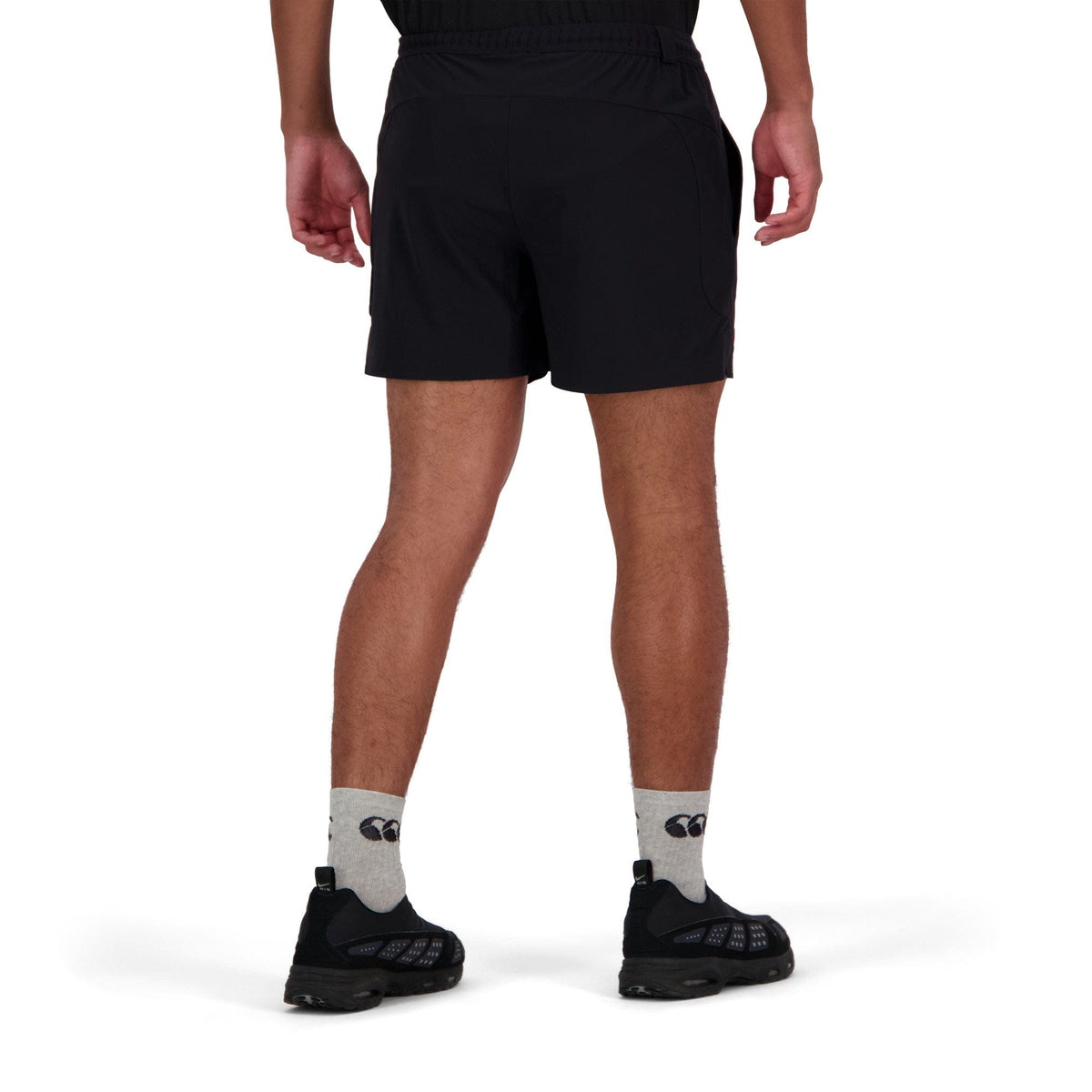 A person stands facing away, wearing Canterbury Everest 5" Woven Short in black, patterned gray socks, and black shoes against a white background.