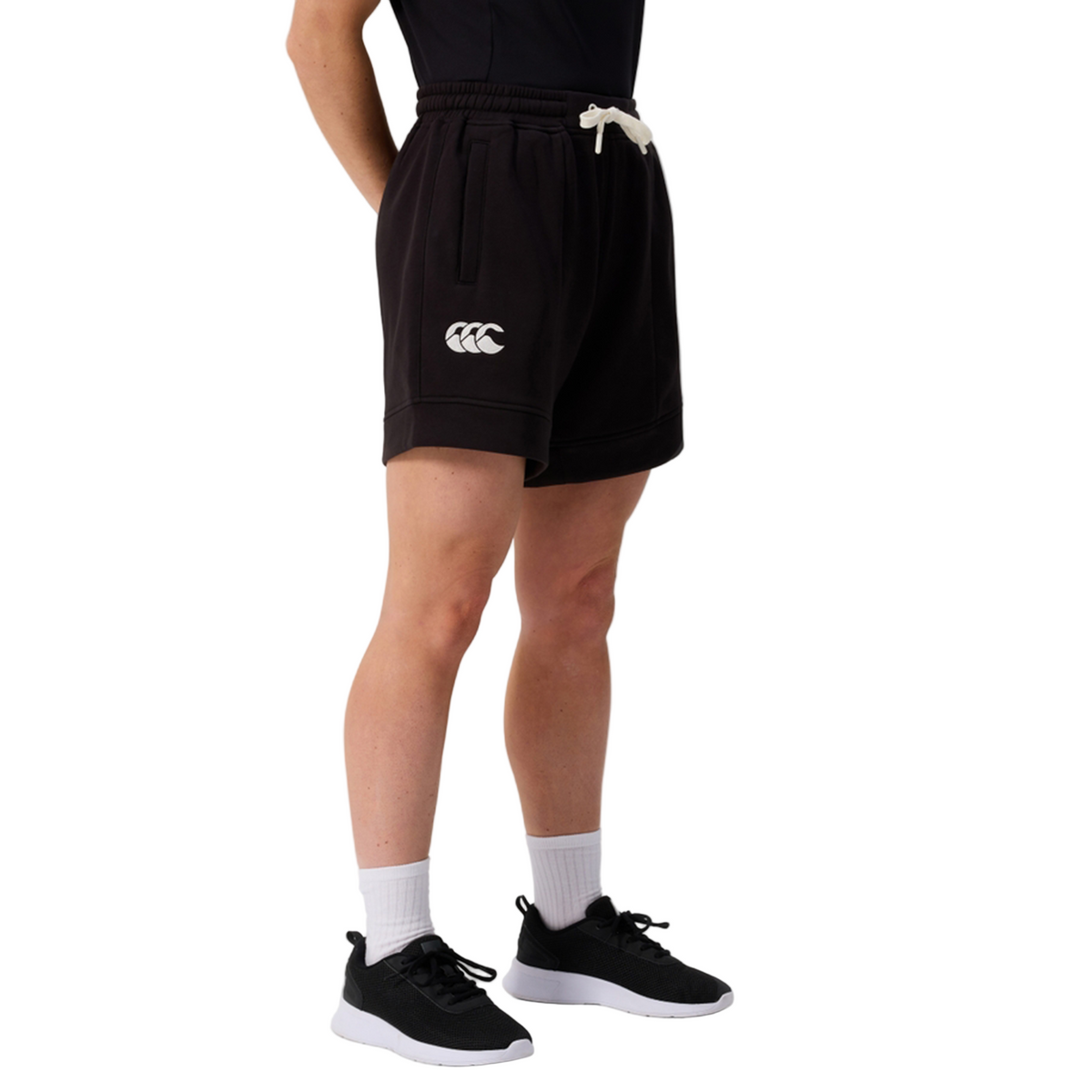 A person wears Canterbury Women&#39;s Ireland 25/26 Knit Shorts with a white logo, white socks, black sneakers, and a black top, standing against a plain light background.