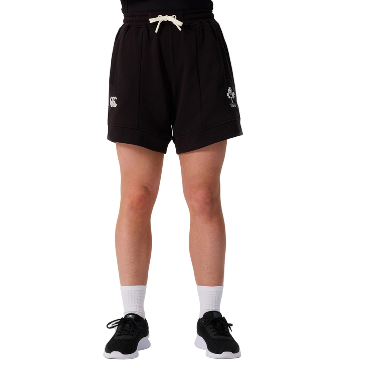 A person stands against a white background, showing only the lower half while wearing Canterbury Women&#39;s Ireland 25/26 Knit Shorts, paired with white socks and black sneakers—highlighting these stylish sports shorts by Canterbury.
