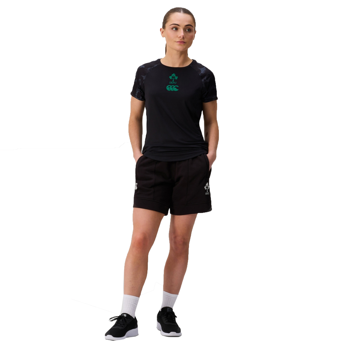 A person stands with hands in pockets, wearing a black athletic t-shirt, Canterbury Women&#39;s Ireland 25/26 Knit Short, white socks, and black sneakers against a white background.