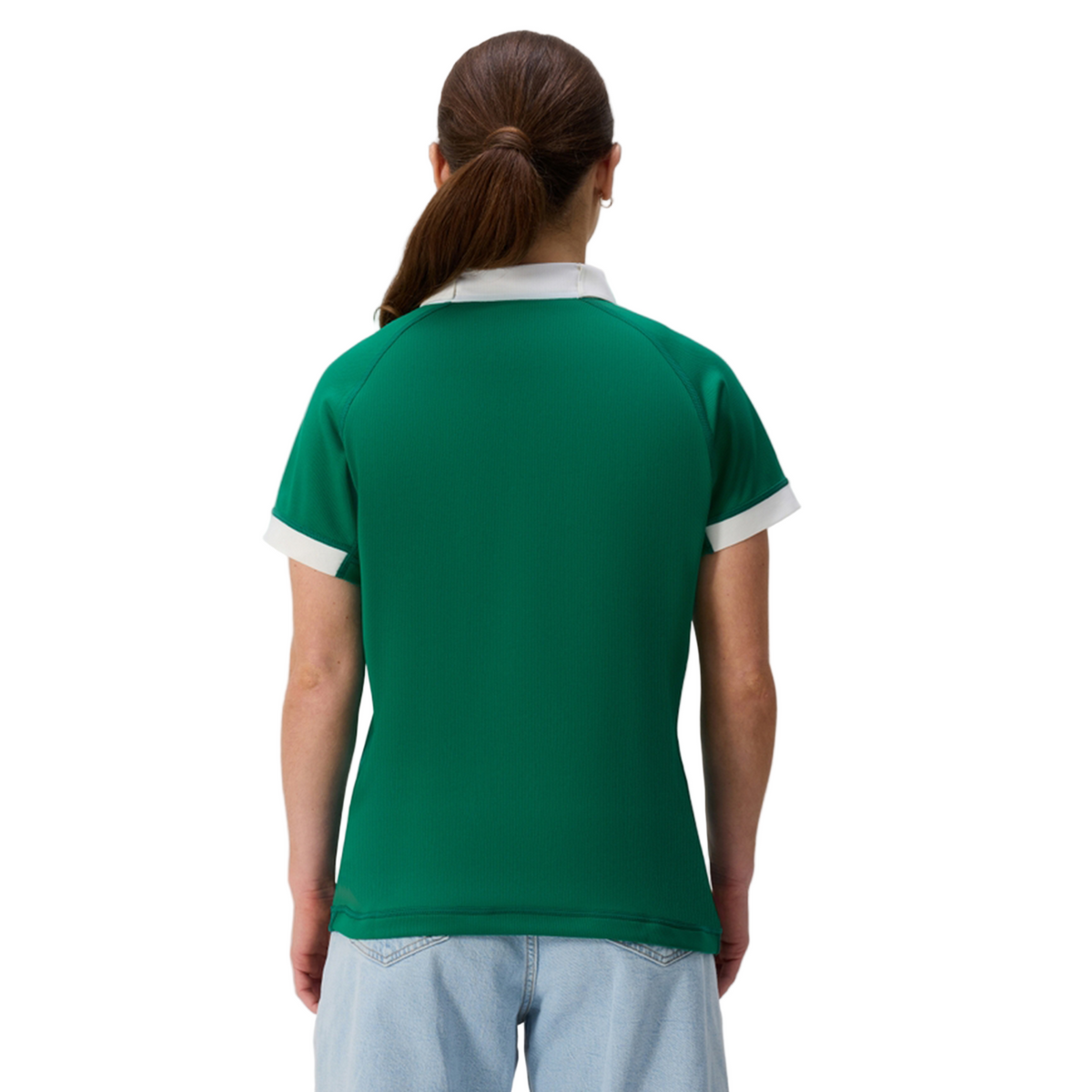 A woman with long hair in a ponytail is seen from the back wearing the Canterbury Women&#39;s Ireland 25/26 Home Pro Jersey with white trim and light blue jeans, standing against a plain background.