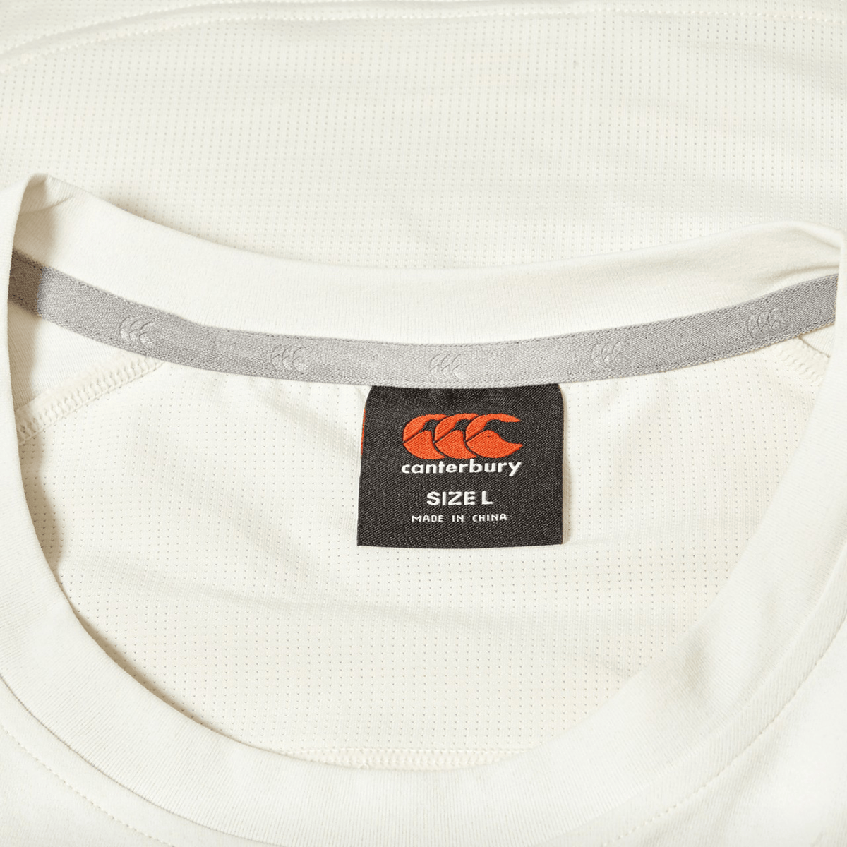 Close-up of a white Canterbury Everest Dart Tech T-Shirt featuring a black and orange Canterbury label indicating size L and made in China.