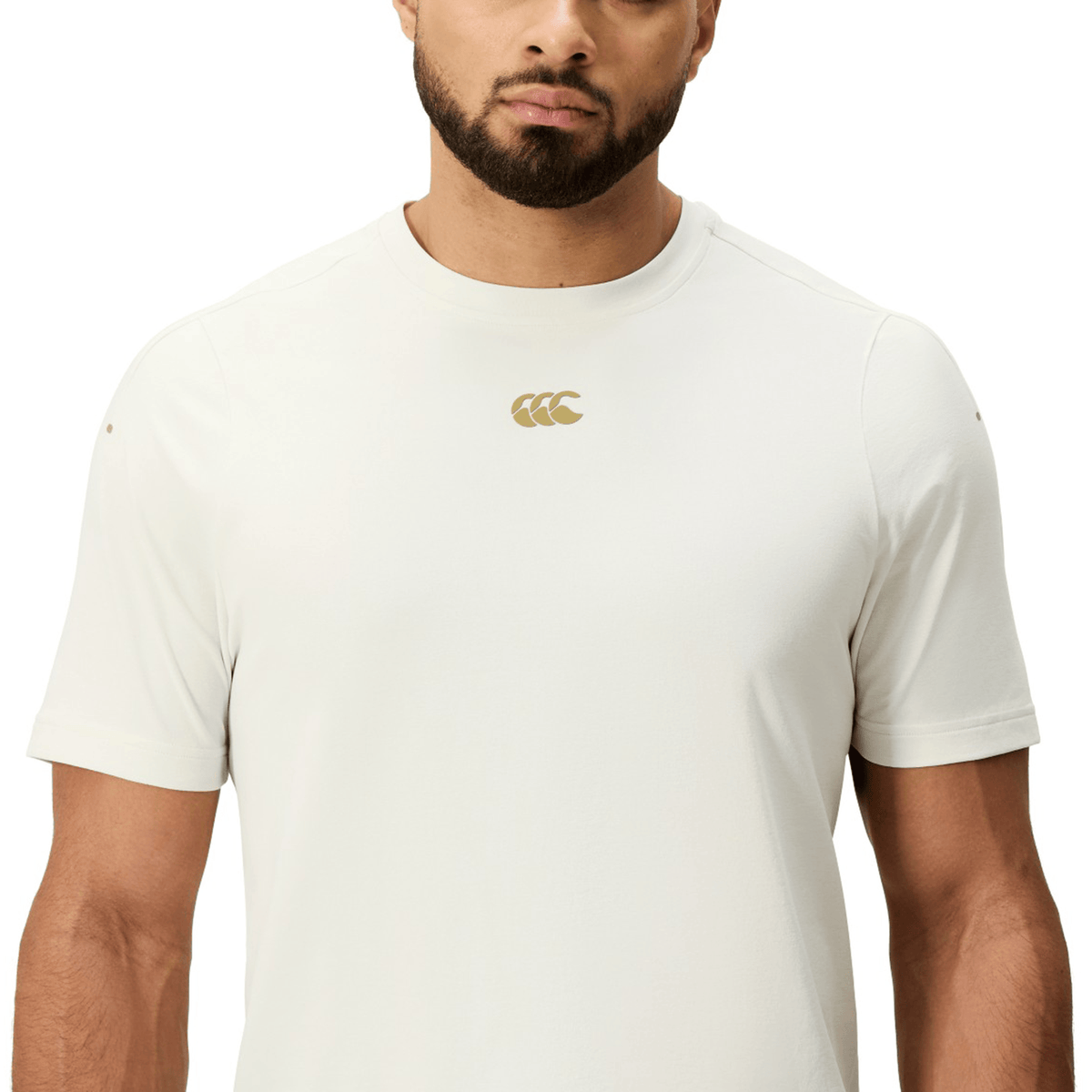 A man wears the Canterbury Everest Dart Tech T-Shirt by Canterbury, a fitted off-white quick-drying short-sleeve top with a small gold chest logo, shown from shoulders to waist against a plain background.