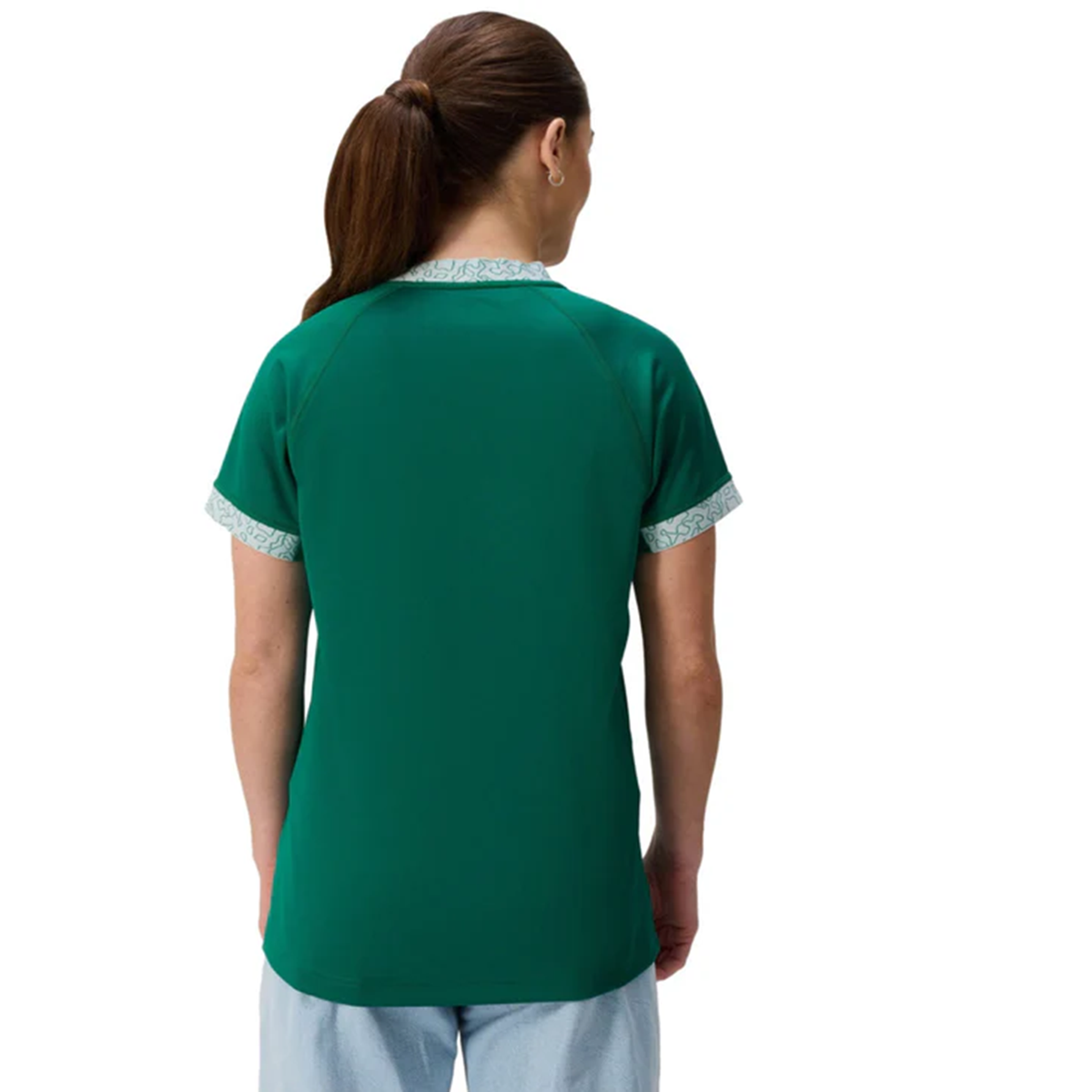 A woman with brown hair in a ponytail wears the Canterbury Women&#39;s Ireland WRWC25 Replica Home Jersey with patterned trim and light blue pants, shown from behind against a white background.