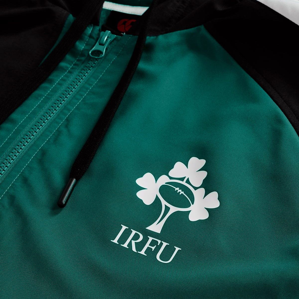 The Ireland 25/26 Legends 1/4 Zip Jacket by Canterbury features a close-up, angled view of its white IRFU logo with a rugby ball and three shamrocks on a green and black design.