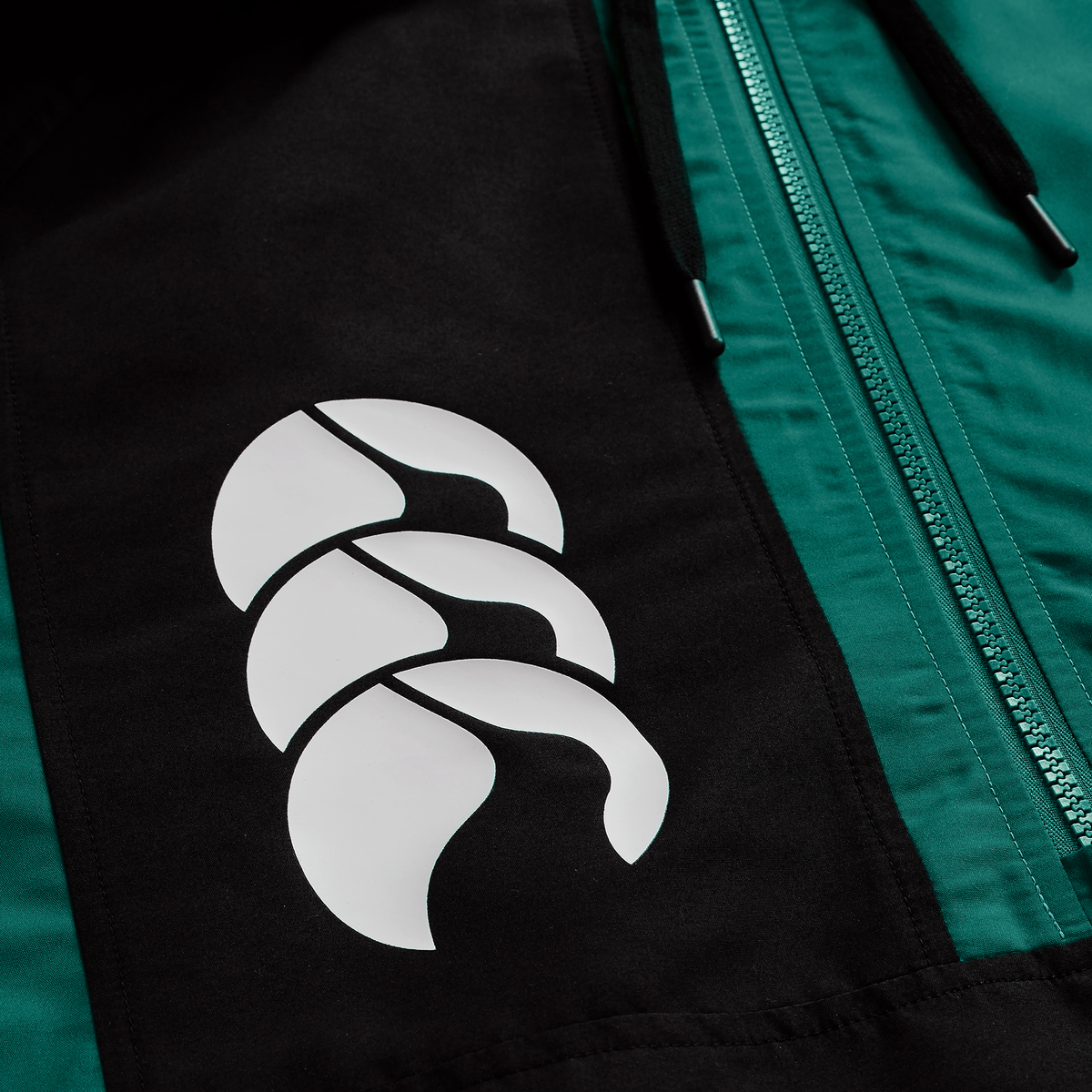 A close-up of the Ireland 25/26 Legends 1/4 Zip Jacket by Canterbury features a white abstract logo and visible green zipper, emphasizing the brand&#39;s stylish quarter-zip design.