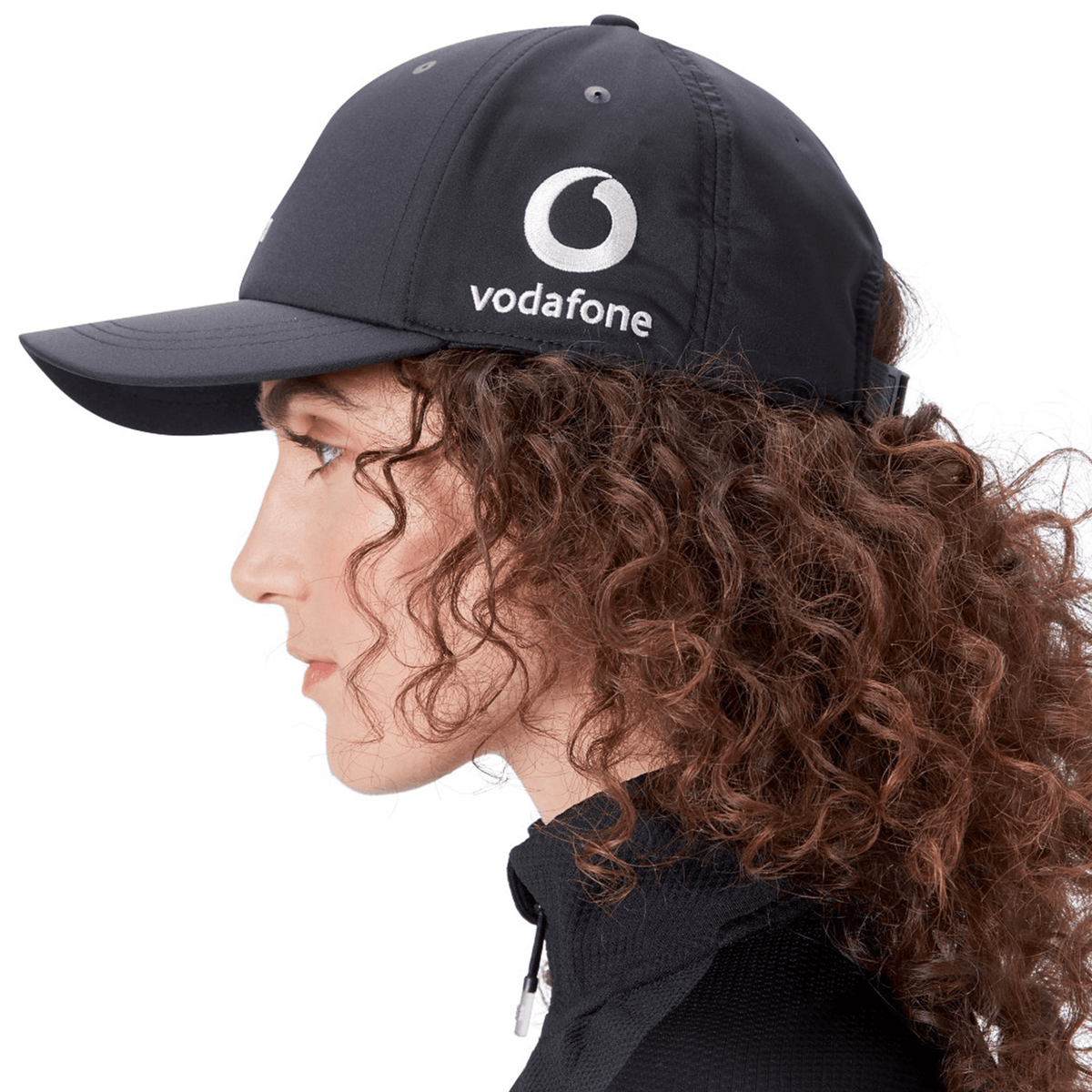Individual with curly hair wears the Ireland 25/26 Tech Logo Cap by Canterbury, a black adjustable sports cap, and a black zip-up top, shown in profile facing left.