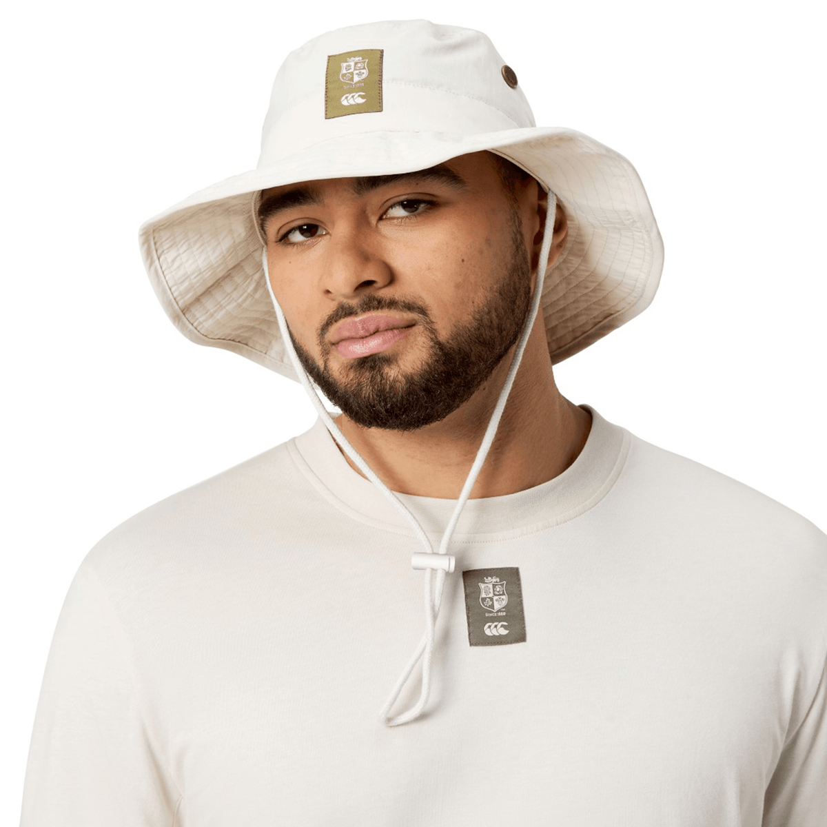 A person wearing the British &amp; Irish Lions 2025 Outback Wide Brim Bucket Hat by Canterbury, made of white quick-dry ripstop fabric, and a long-sleeve shirt with a logo tag, looking at the camera.