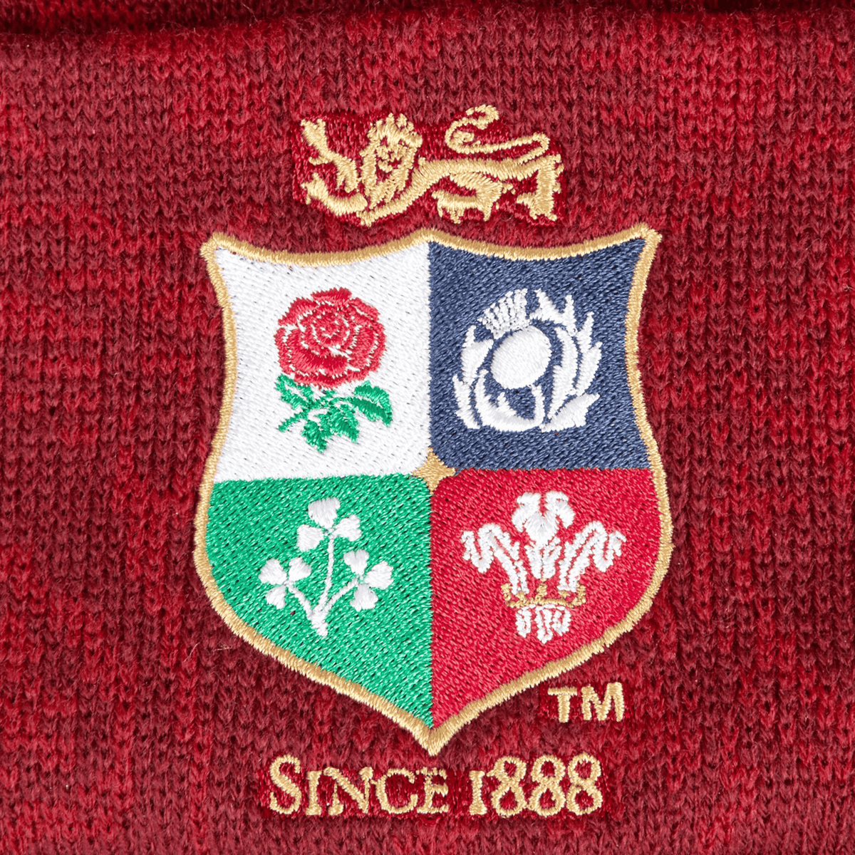 A close-up of the red dahlia-colored British &amp; Irish Lions 2025 Pom-Pom Beanie by Canterbury showcases an embroidered lion logo above a shield decorated with four emblems: a rose, thistle, shamrock, and feathers. Beneath is the text "Since 1888," highlighting the longstanding heritage of the British &amp; Irish Lions.