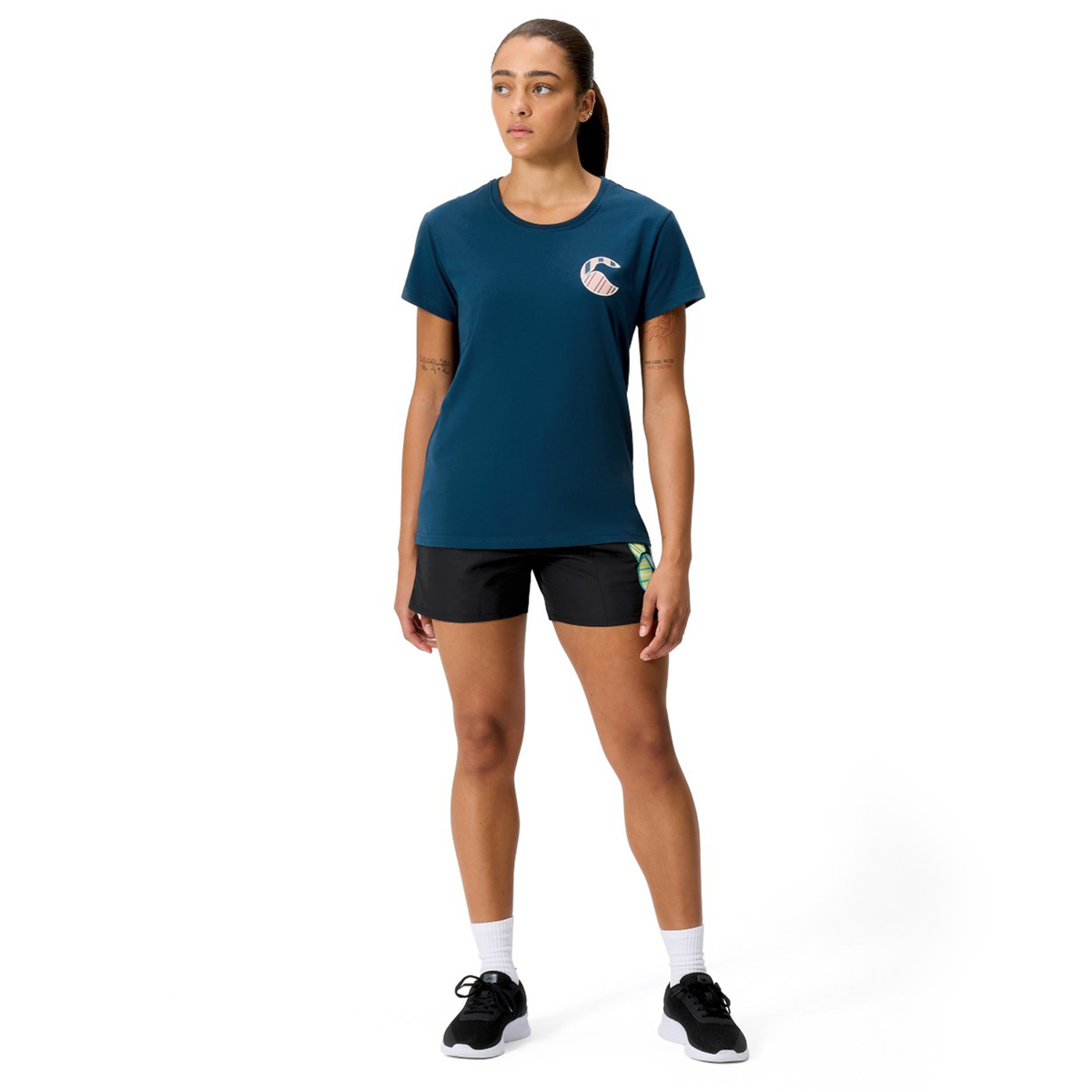 A person stands facing forward in a Canterbury Women&#39;s Uglies Logo T-Shirt, paired with black shorts, white socks, and black sneakers against a plain white background.