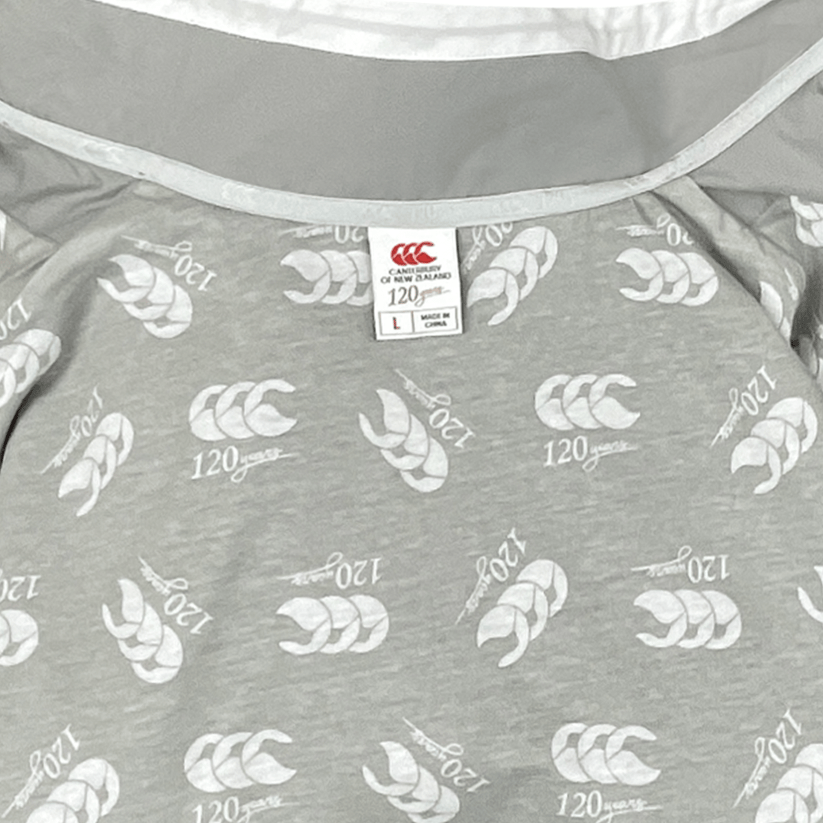 Close-up of a gray Canterbury Legends Woven Track Jacket featuring a repeated white feather pattern and the Canterbury logo, showing the size label inside the collar.